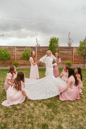Bridesmaids in pink dresses help arrange a bride's wedding gown train outdoors.