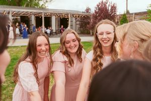 Group in matching pink bridesmaid dresses share a candid laughing moment at outdoor wedding venue.