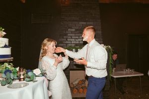 A bride and groom playfully feed each other cake during their wedding reception by a brick fireplace.