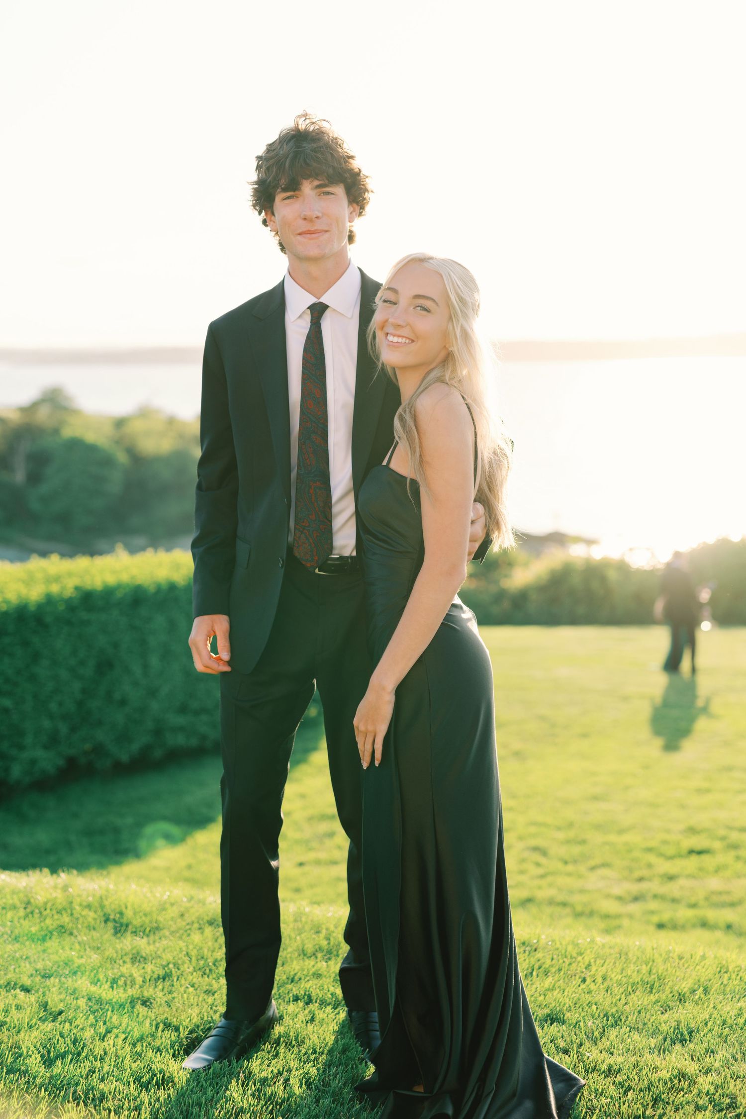 A formal couple in evening attire poses together on a grassy lawn at sunset with water and greenery in the background.