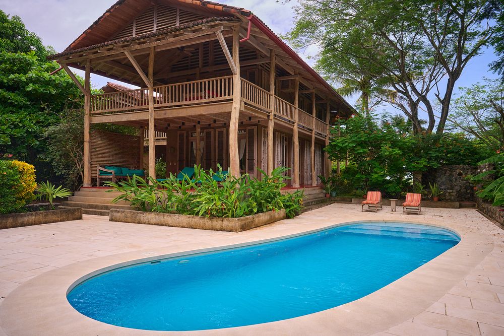 Luxury tropical villa with wooden deck and turquoise swimming pool surrounded by lush green landscaping in paradise setting.