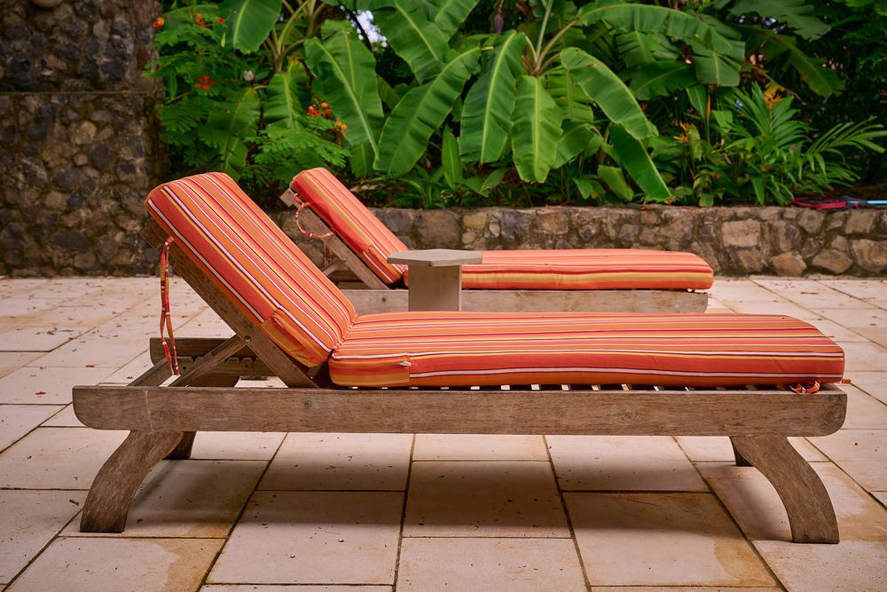 Two orange cushioned wooden chaise lounge chairs sit on a stone patio surrounded by tropical landscaping.