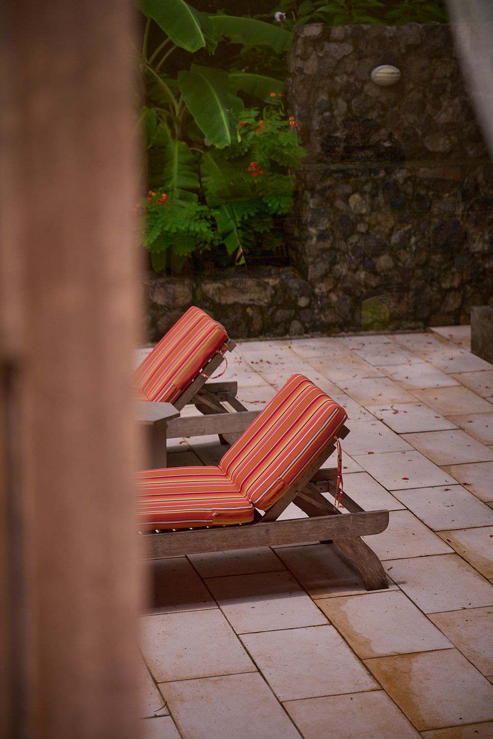 Coral-colored cushions on wooden loungers set against a rustic stone wall and tropical garden backdrop.