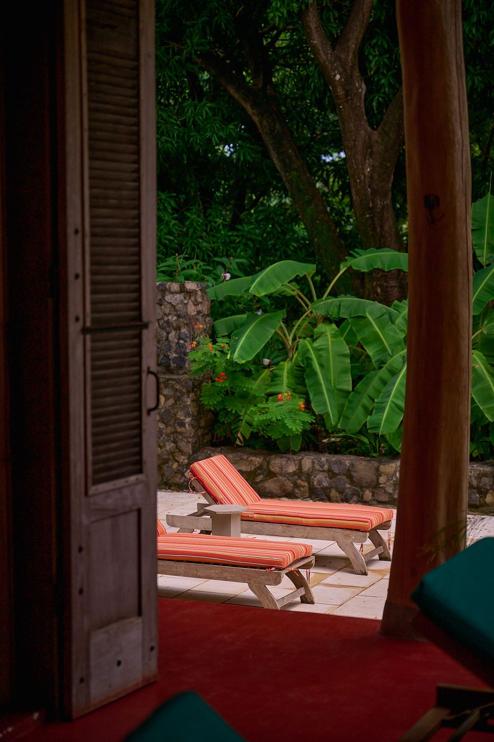 Two coral lounge chairs on a patio overlooking lush tropical gardens through an open doorway with wooden shutters.