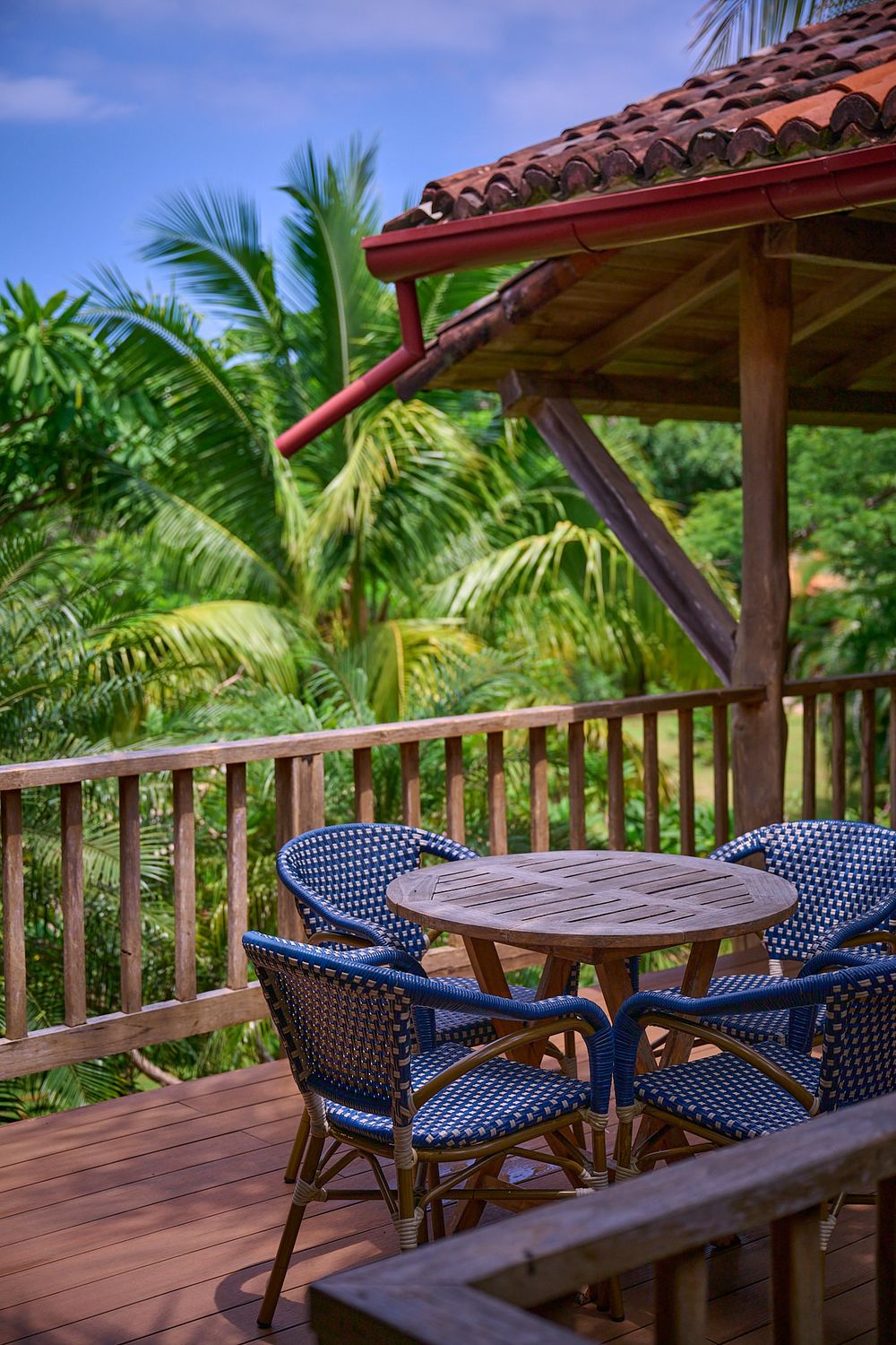Panoramic view of a tropical restaurant deck with blue bistro chairs and wooden tables surrounded by lush palm trees.