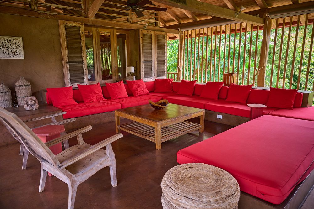 Outdoor covered patio with bright red cushioned sectional seating and wooden coffee table in tropical setting.
