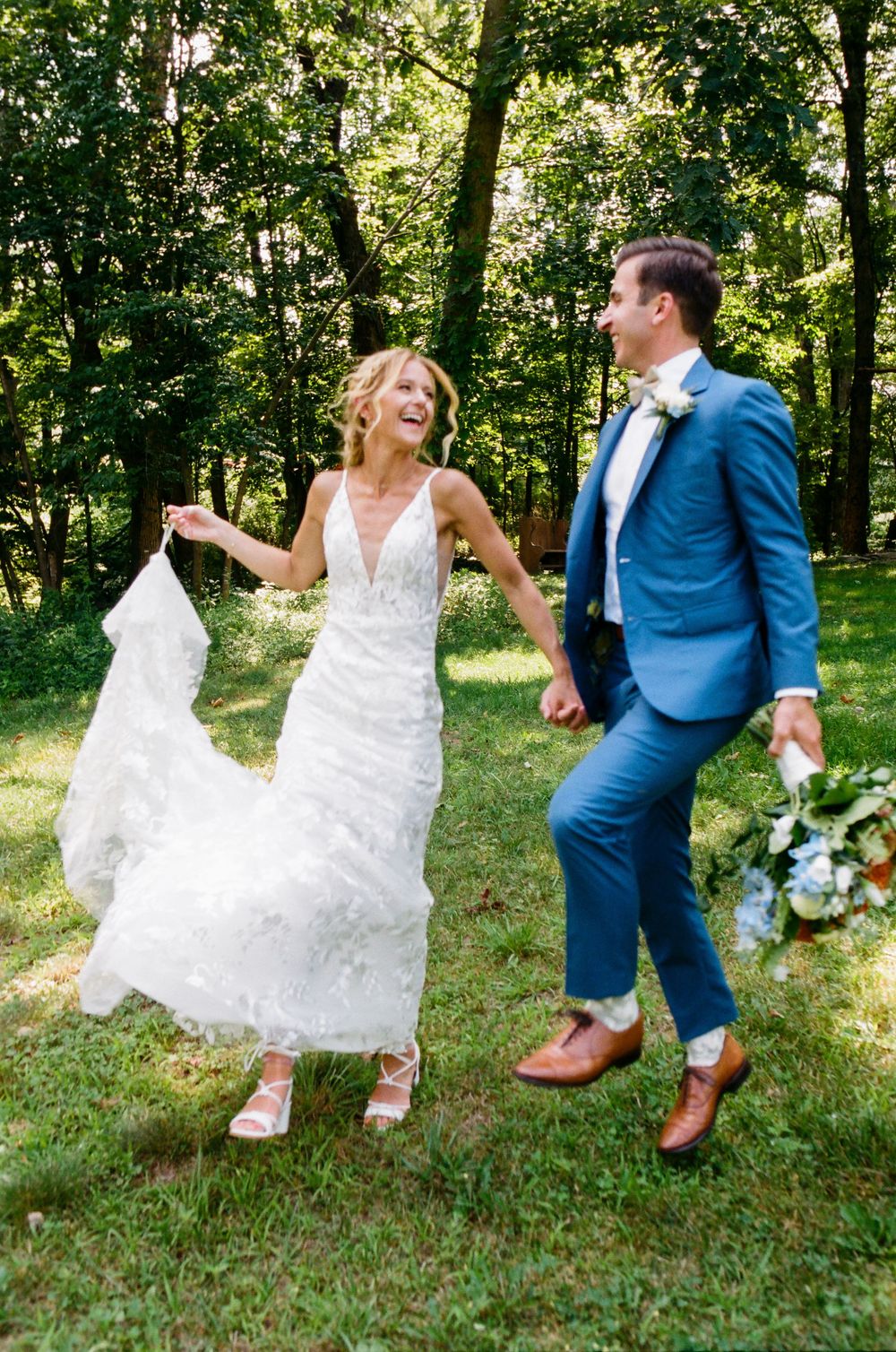 Playful wedding couple dancing on grass in elegant dress and blue suit.
