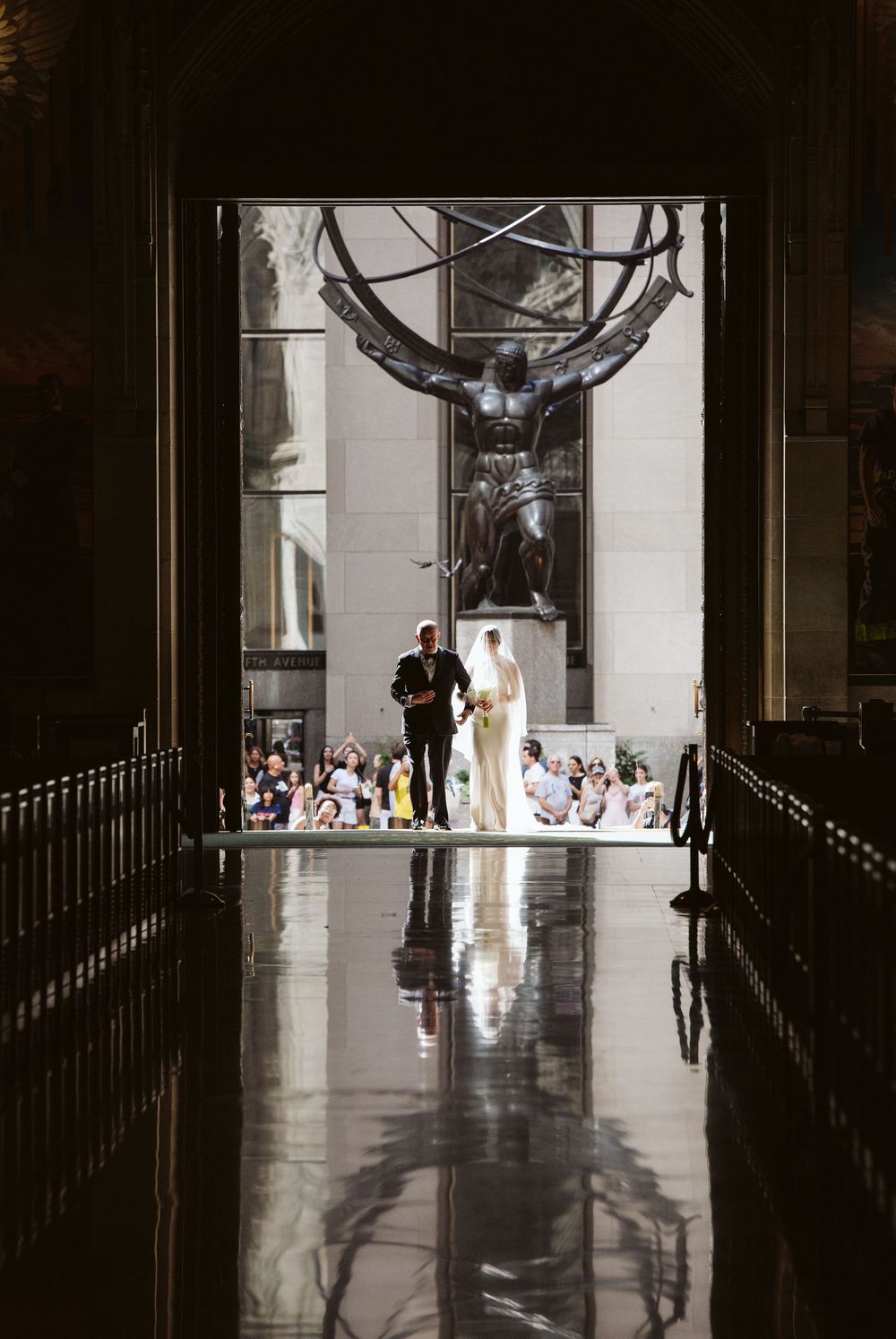 Bride and groom stand before the iconic Atlas statue at Rockefeller Center, reflected on a gleaming floor.