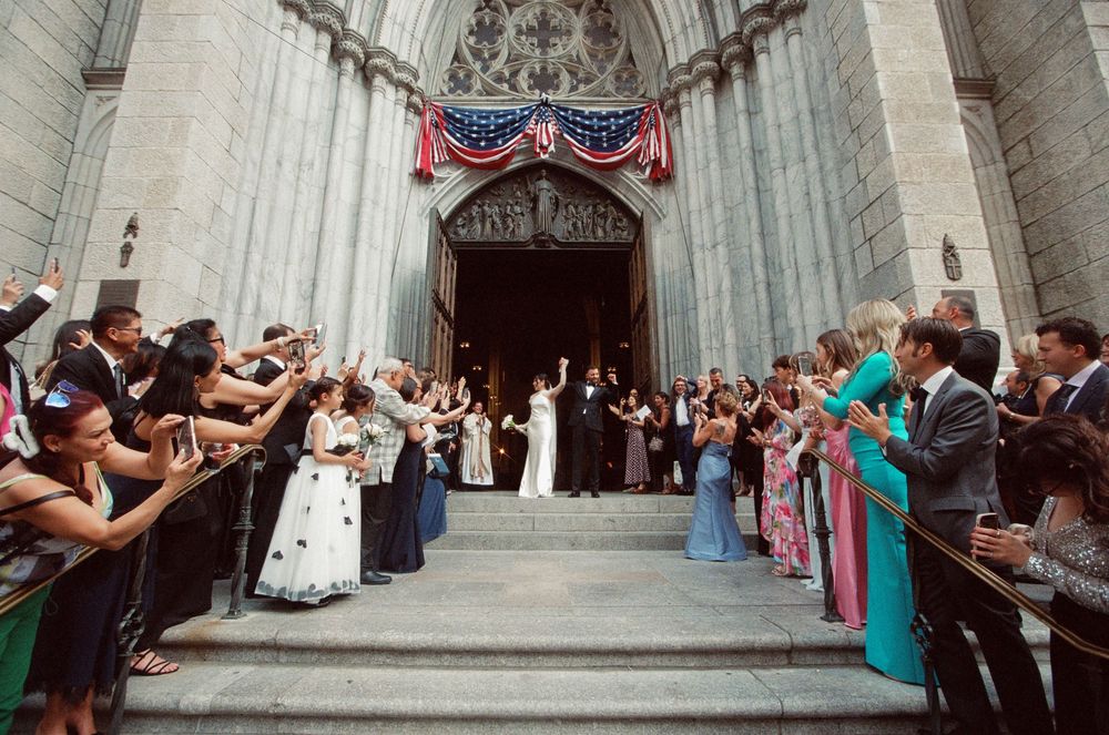 Wedding couple exits St. Patrick's Cathedral NYC as guests line steps cheering with American flag banner.