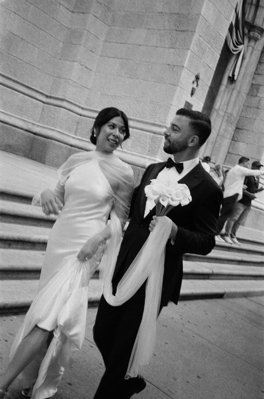 Black and white photo of a couple in wedding attire, bride in white gown and groom in tuxedo, walking happily outside.