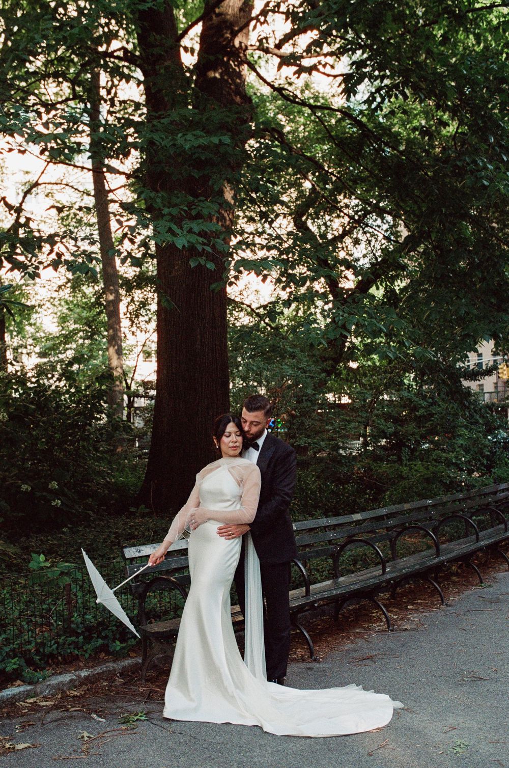 Bride in white gown and groom in tuxedo embrace in Central Park, surrounded by lush green trees.