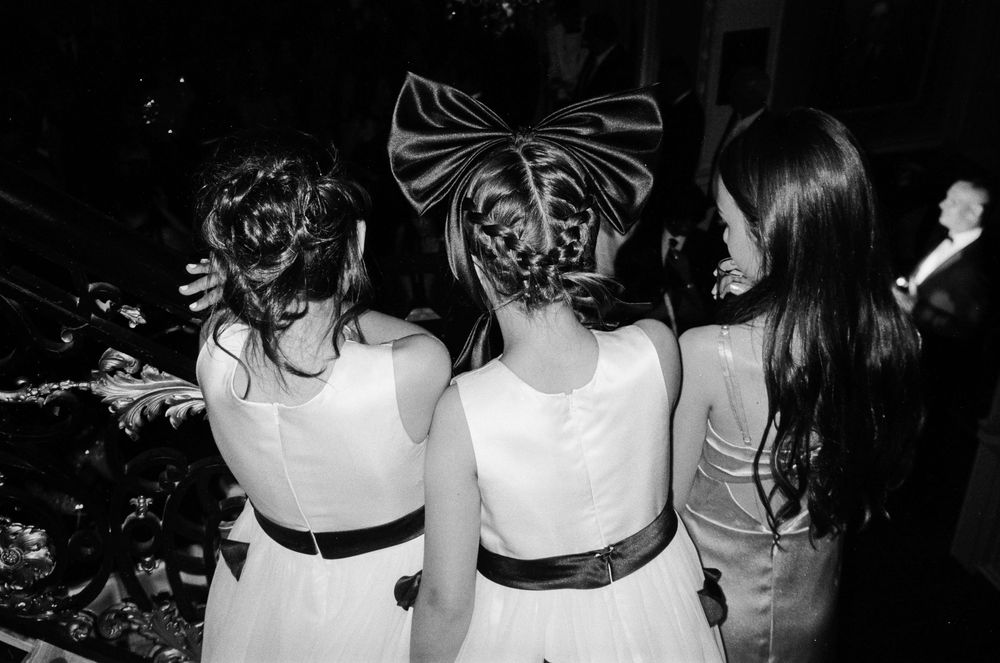 Three young flower girls in white dresses with black sashes, backs turned, one wearing a large satin bow in her hair.