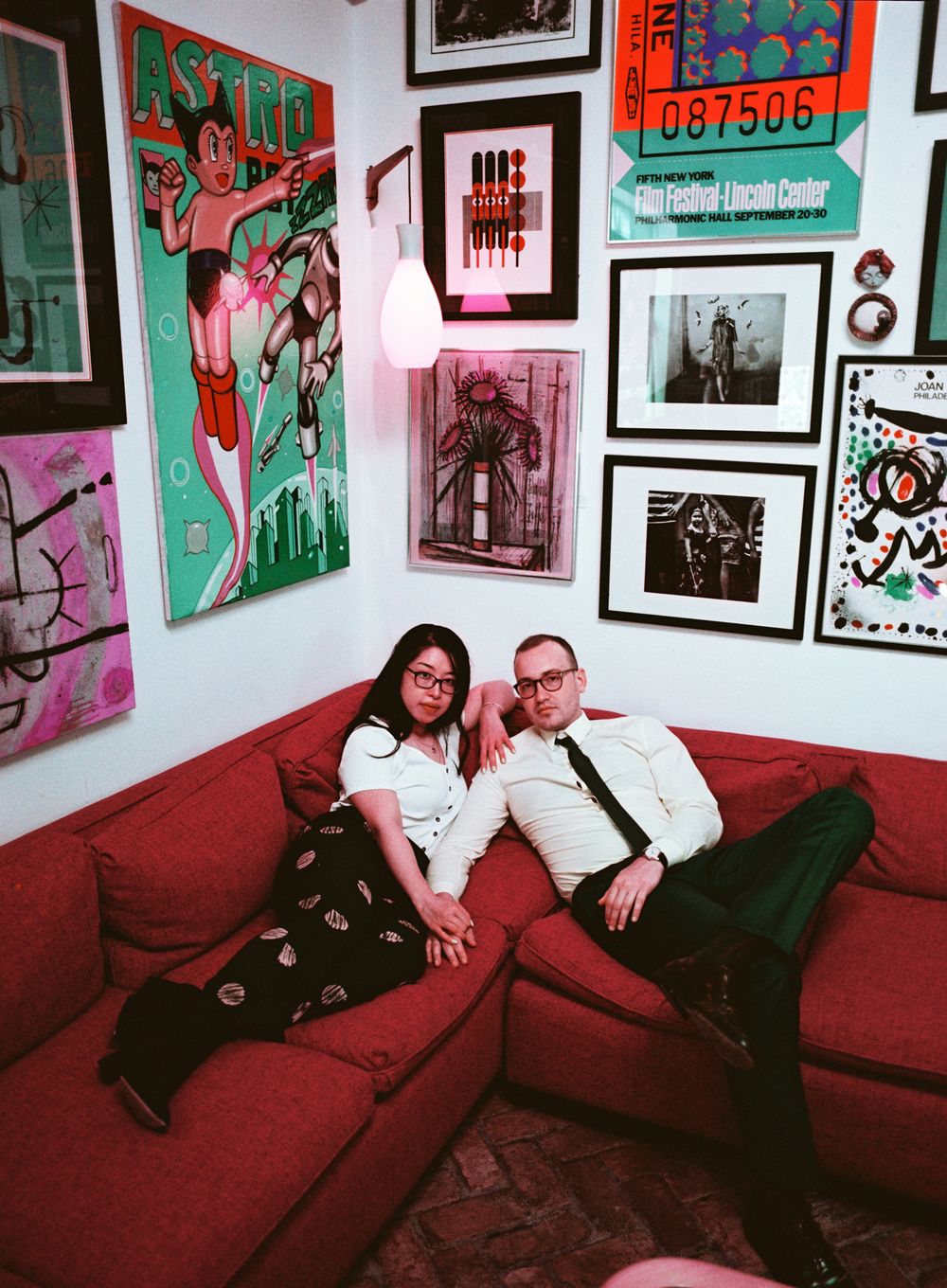 Couple relaxing on red sofa surrounded by eclectic gallery wall with colorful posters and framed artwork.