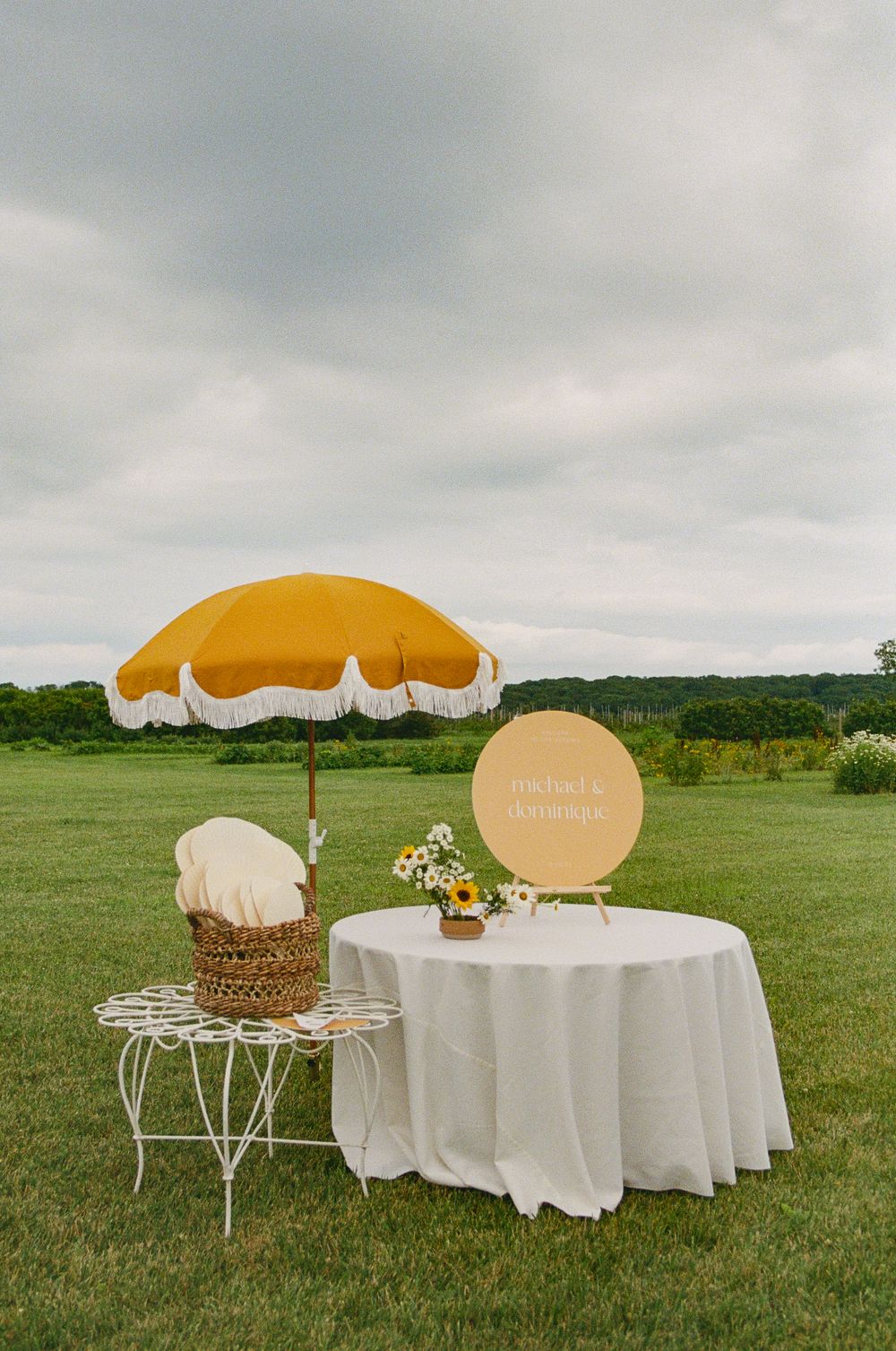 Outdoor wedding setup with yellow umbrella, white tablecloth, and floral centerpiece on a green lawn.