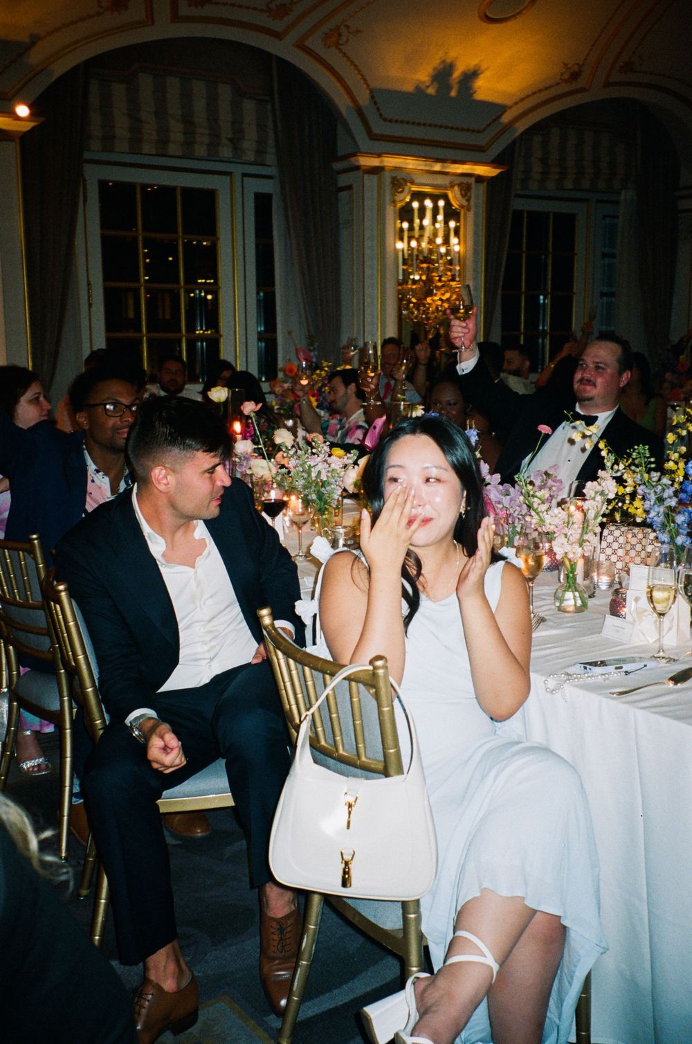 Wedding reception guests celebrating at decorated dining tables under elegant chandeliers.