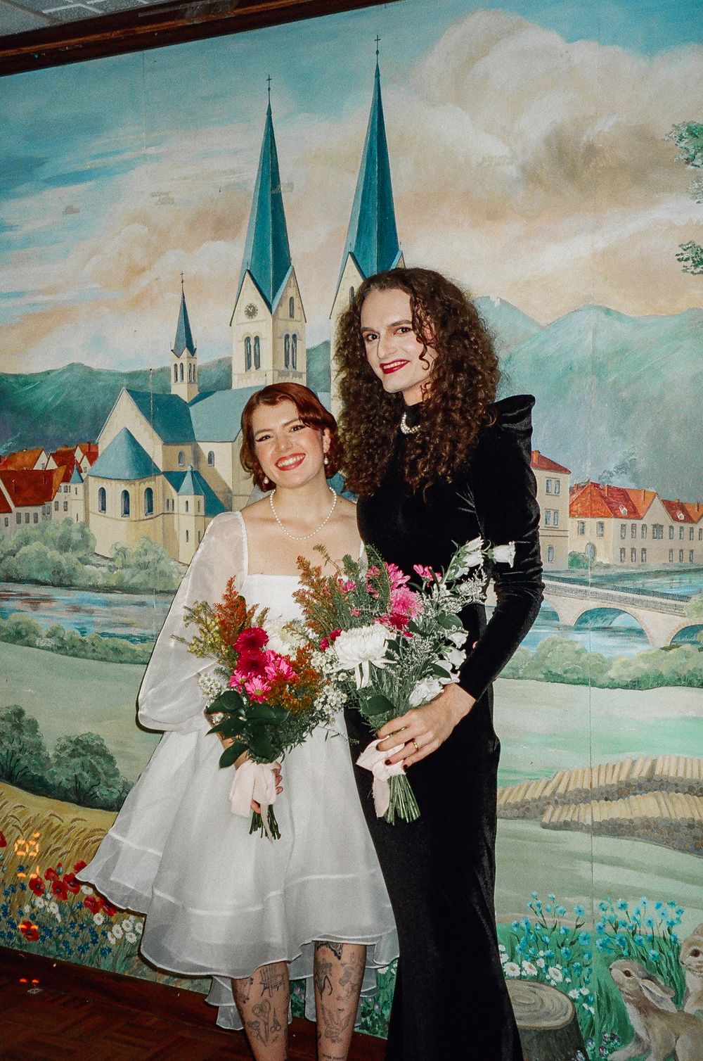 Couple posing with bouquets against a painted mural backdrop featuring a church and landscape scene.