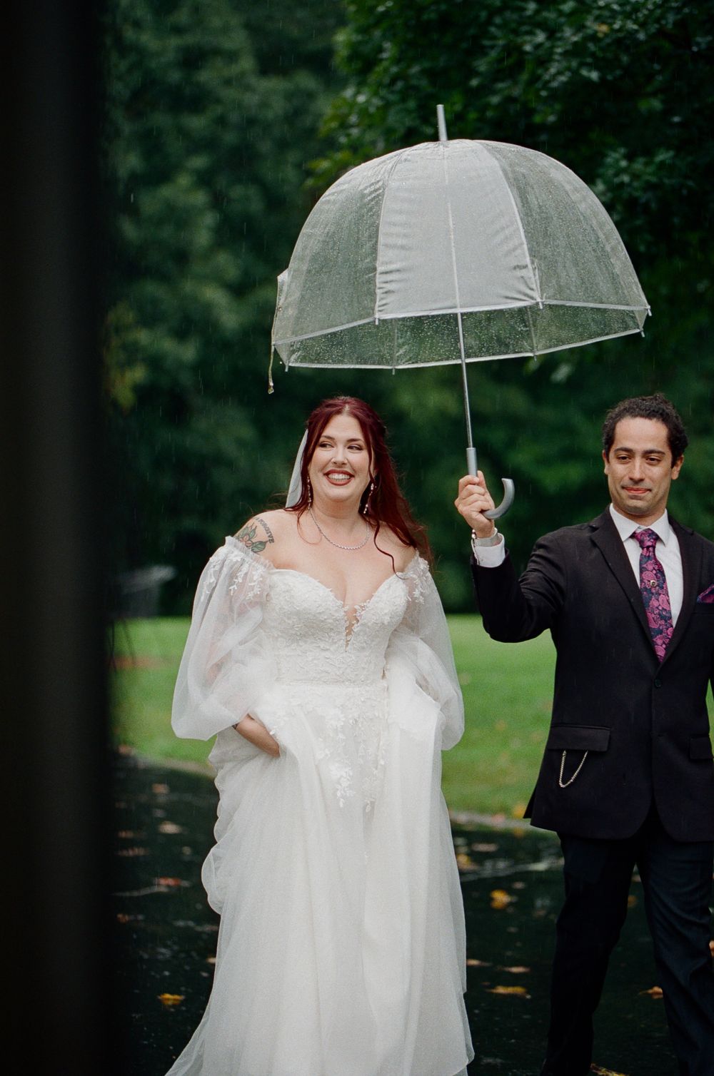 Smiling bride in a white lace wedding gown walks outdoors in the rain as a groom in a dark suit holds a clear umbrella over her.
