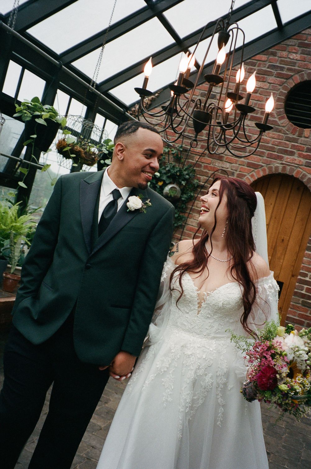 Smiling bride in a lace gown with bouquet holds hands with groom in a dark green suit inside a greenhouse venue with a candle chandelier.