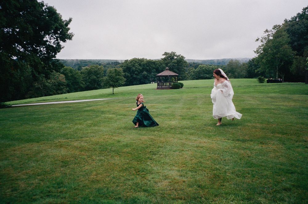 Bride in a white gown and a young girl in a green dress run across a wide grassy lawn toward a gazebo under an overcast sky.