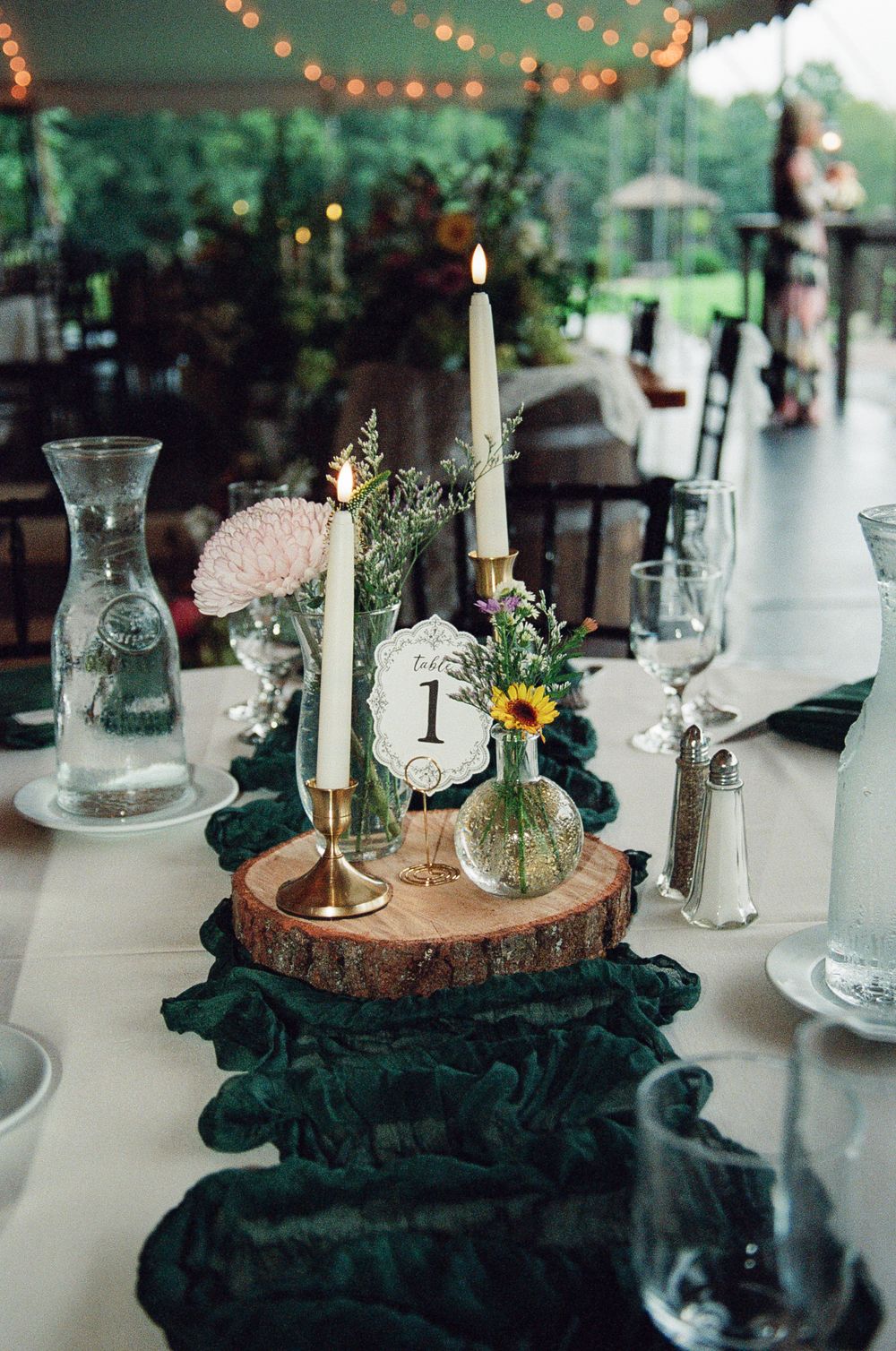 Rustic wedding table with table number 1, taper candles on a wood slice, sunflower bud vase and green velvet runner under string lights.