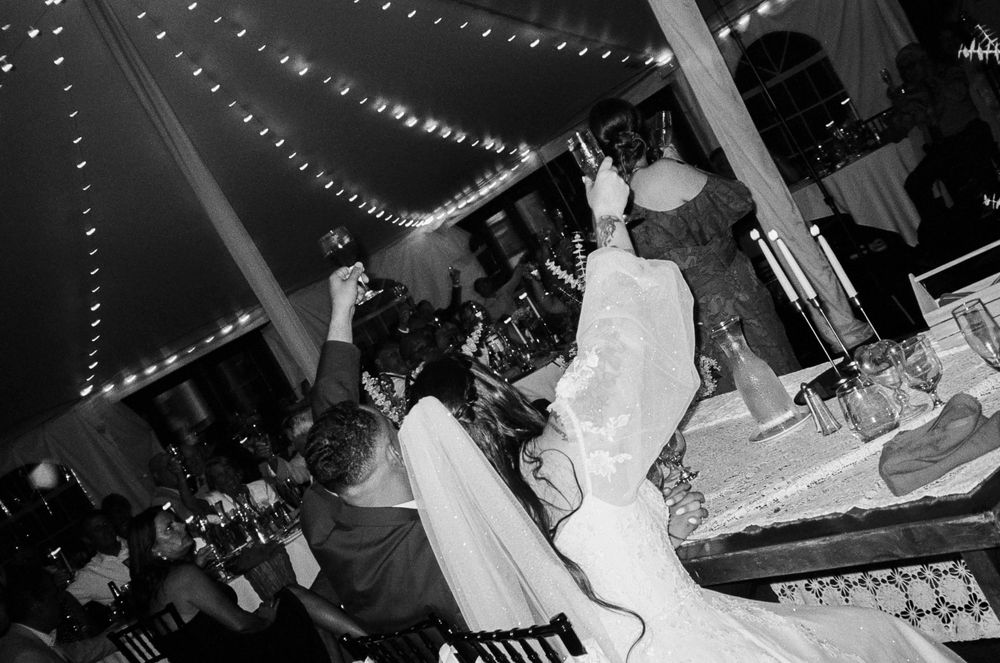 Black and white photo of a bride in a wedding gown raising her arm to cheer with guests at a tented reception with string lights.
