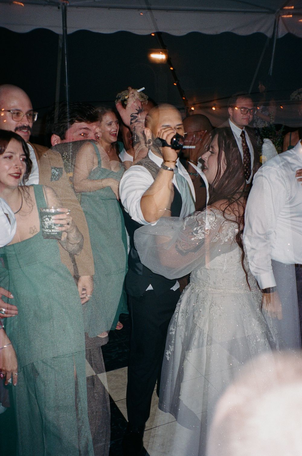 Wedding guests in green dresses and formal attire dance and cheer under a tent as a man sings into a microphone beside the bride.