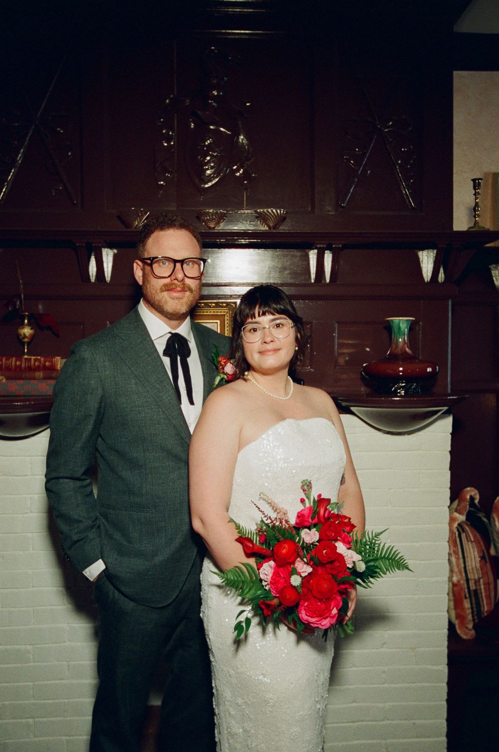 Bride holding red bouquet stands with groom in green suit before dark wood mantle at wedding venue.
