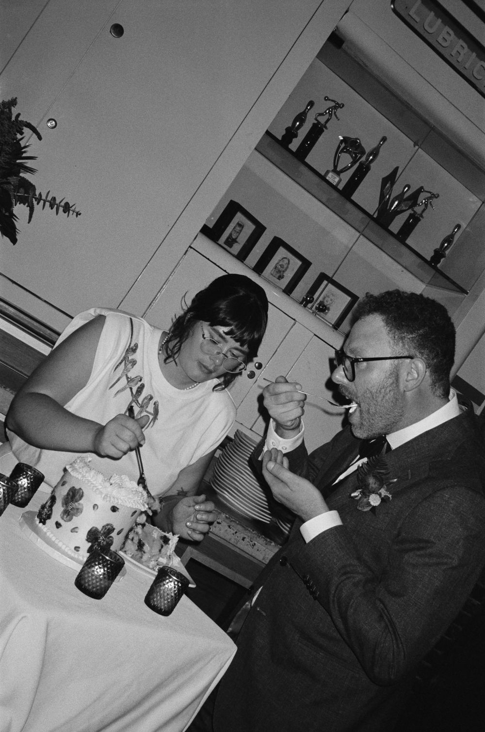 Black and white photo of a couple cutting wedding cake, bride feeding groom at indoor reception celebration.