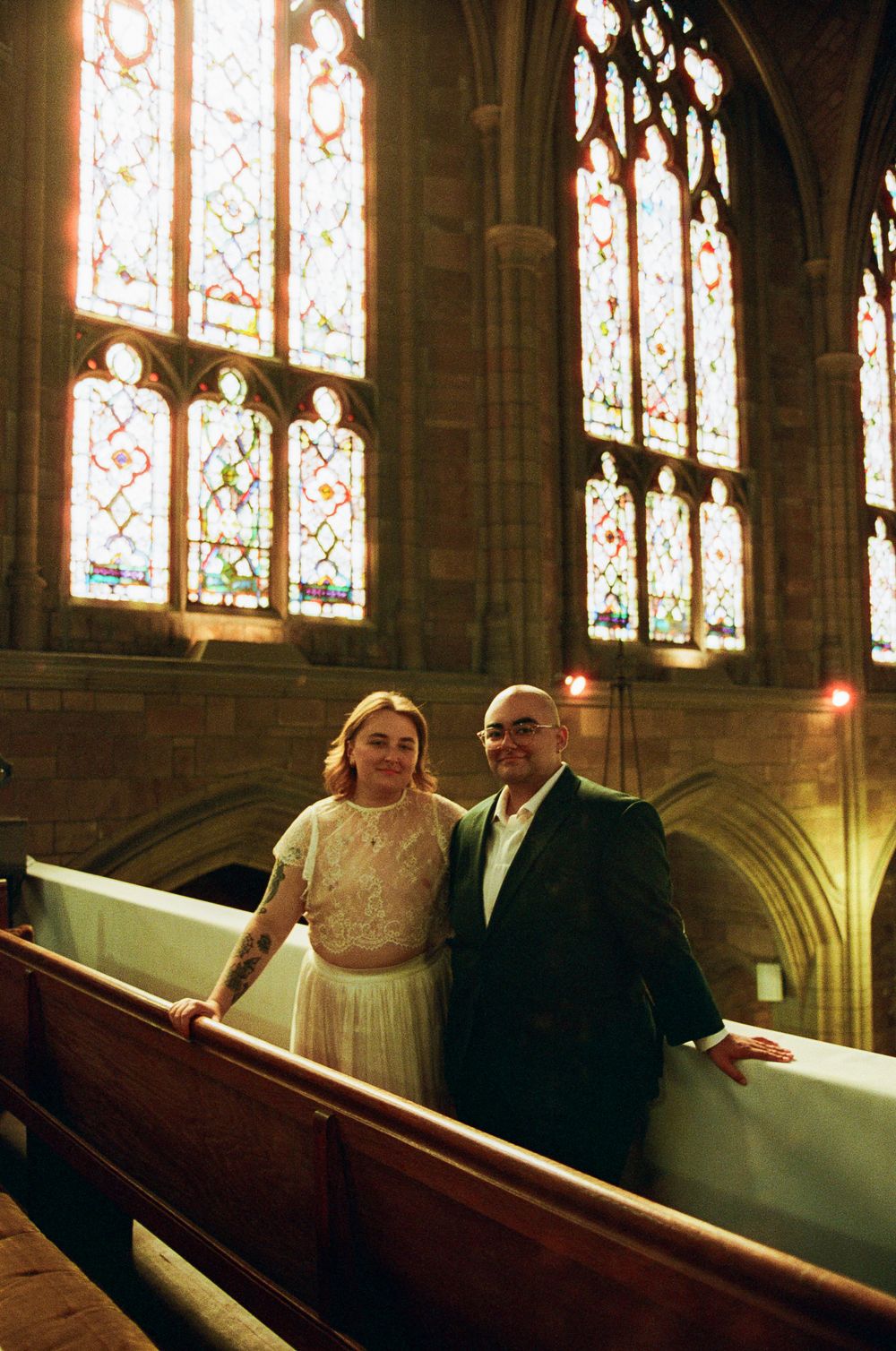 A smiling couple in formal attire stands arm in arm beside a wooden pew inside a Gothic church, glowing stained glass windows behind them.