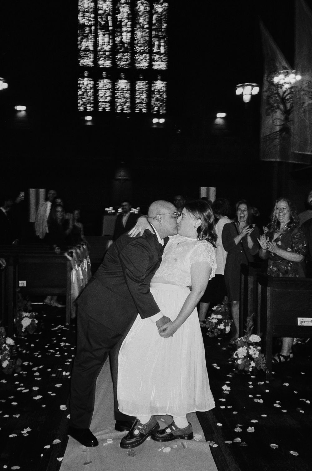 Black and white photo of a bride and groom sharing a kiss in a church aisle strewn with petals as smiling guests applaud behind them.