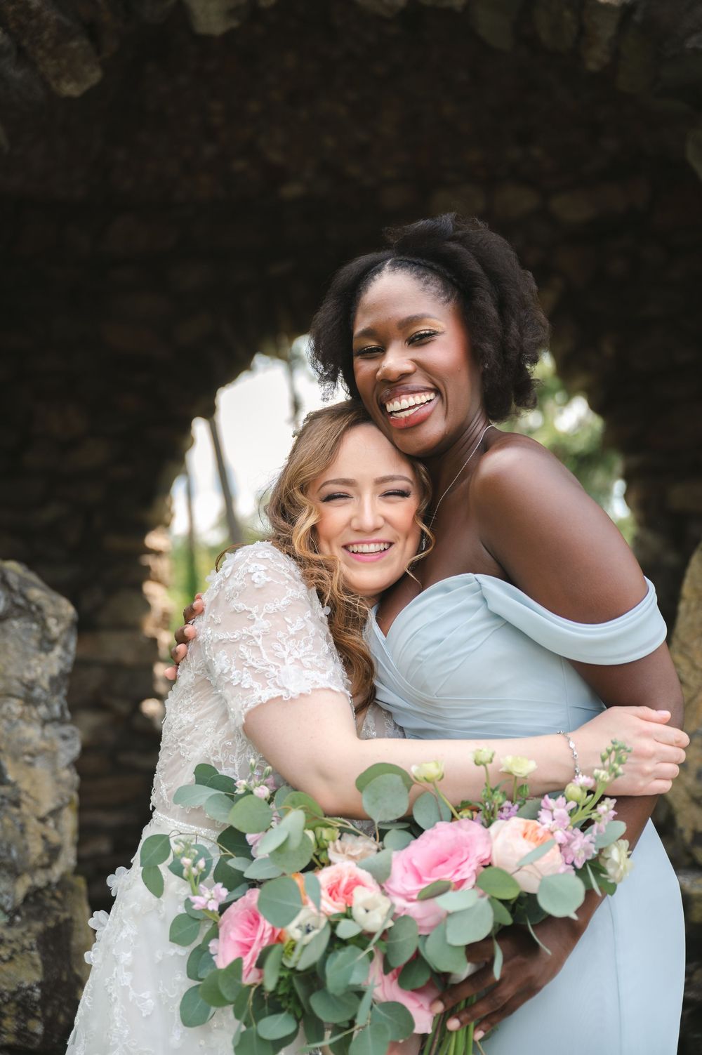 Happy bride and bridesmaid laughing together while holding pink floral bouquet with eucalyptus leaves.