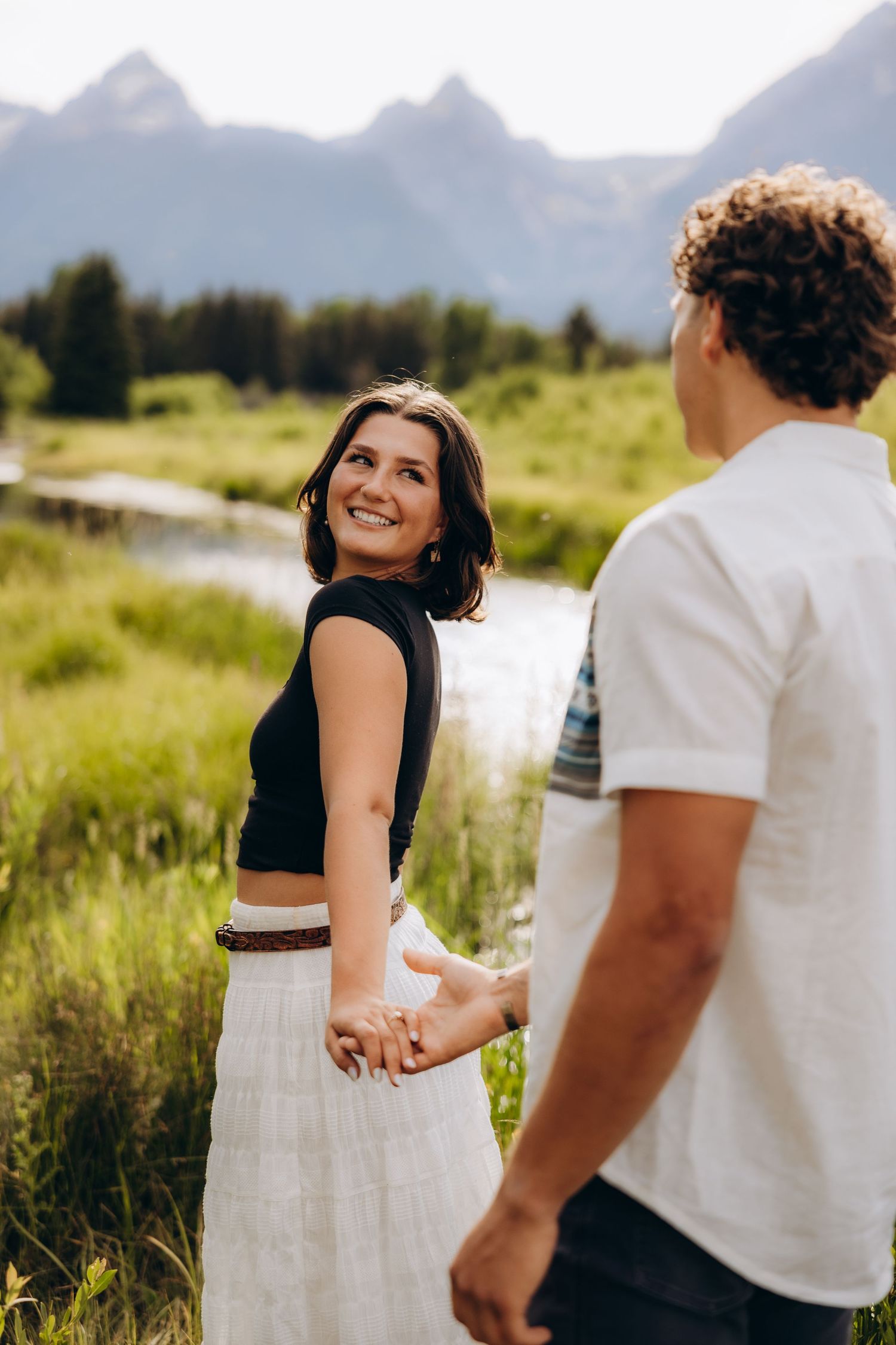 A couple holds hands in a scenic mountain meadow with peaks visible in the background on a sunny day.