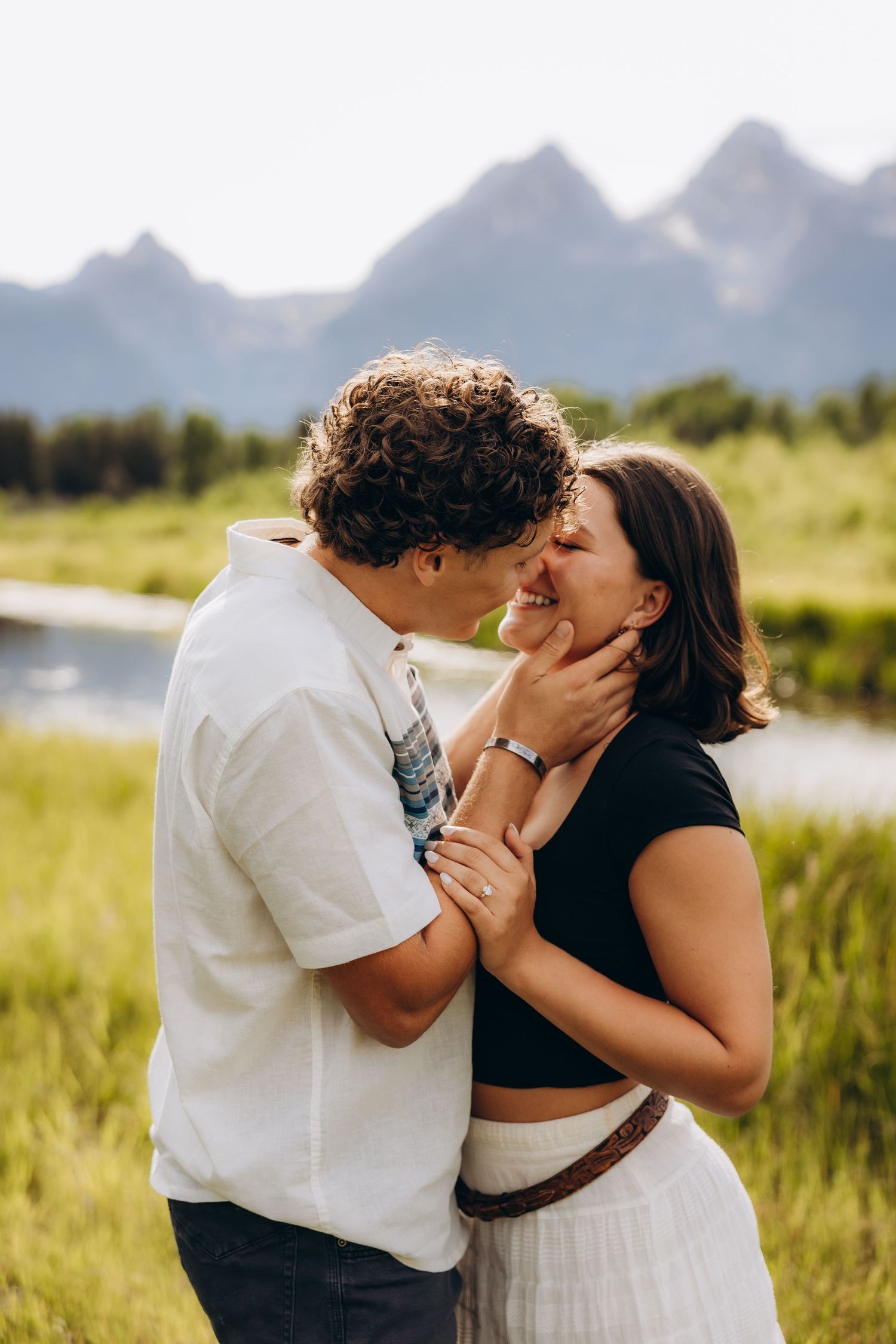 A couple shares a romantic kiss in front of majestic mountain peaks with a river and meadow in the background.