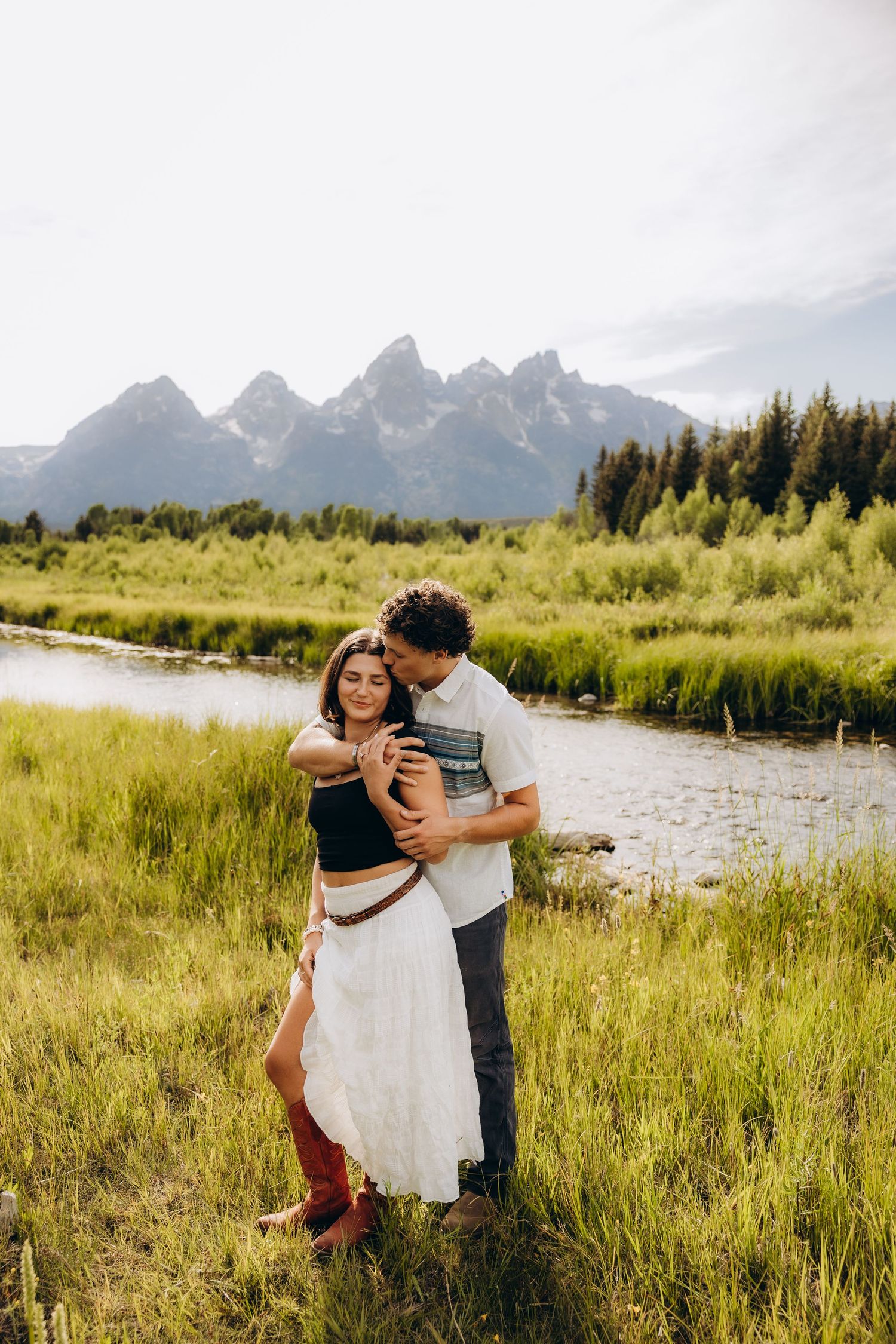 A couple embraces in a grassy meadow with the majestic Grand Teton mountains rising in the background.