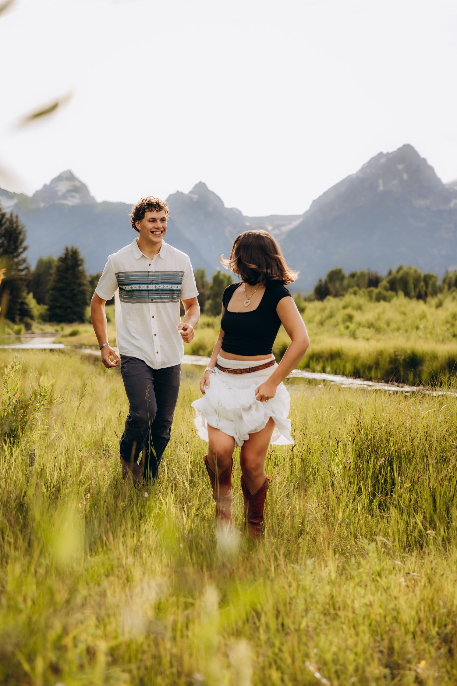 A couple walks together through tall summer grass with majestic mountain peaks rising in the background.