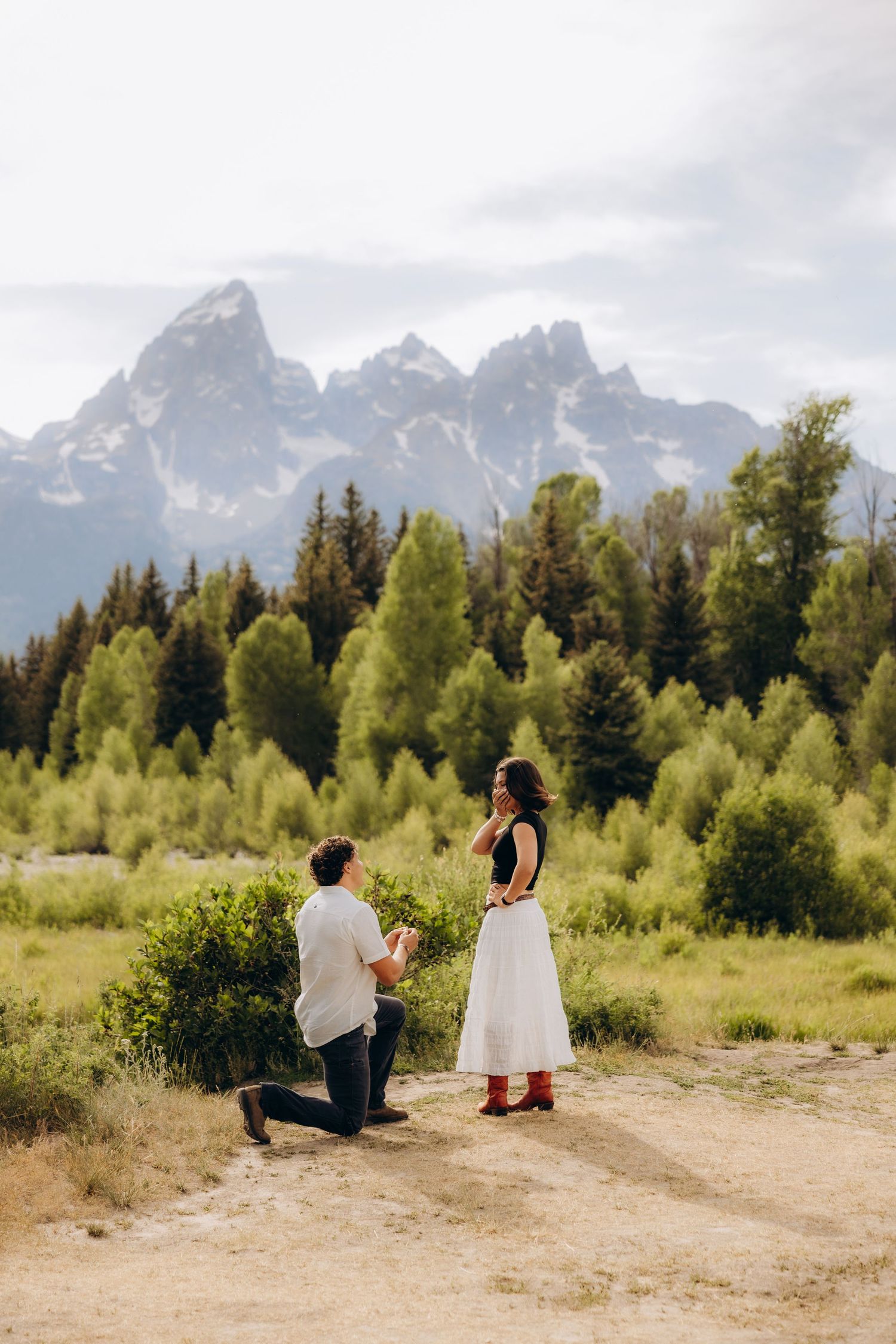 A couple shares a romantic moment in a scenic mountain landscape with evergreen trees and rugged peaks in the background.