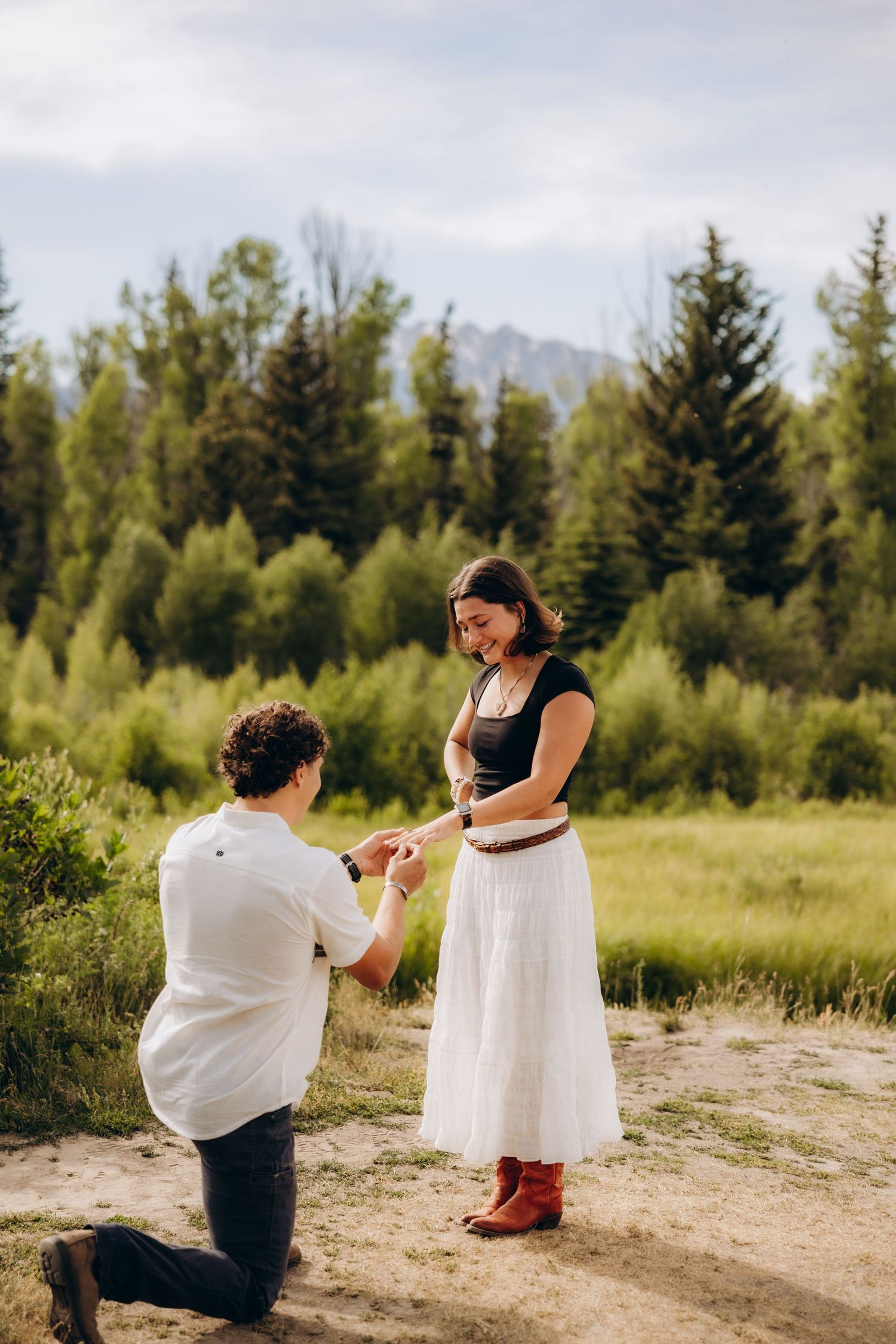 A romantic proposal moment captured in a scenic mountain meadow surrounded by evergreen trees.