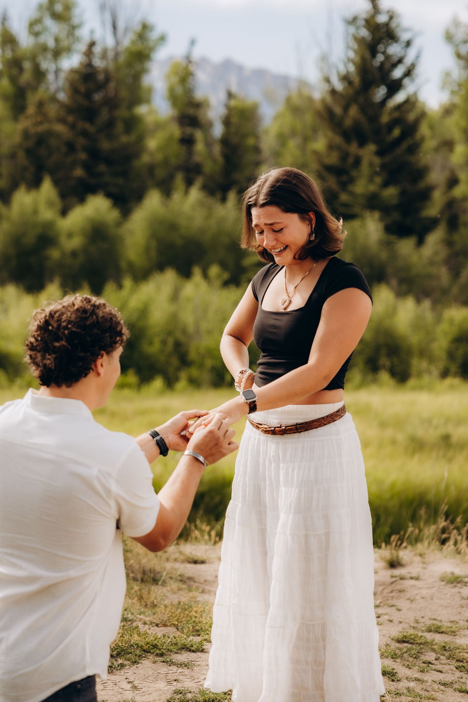 A romantic outdoor proposal sequence against a scenic mountain backdrop with evergreen trees and tall grass.