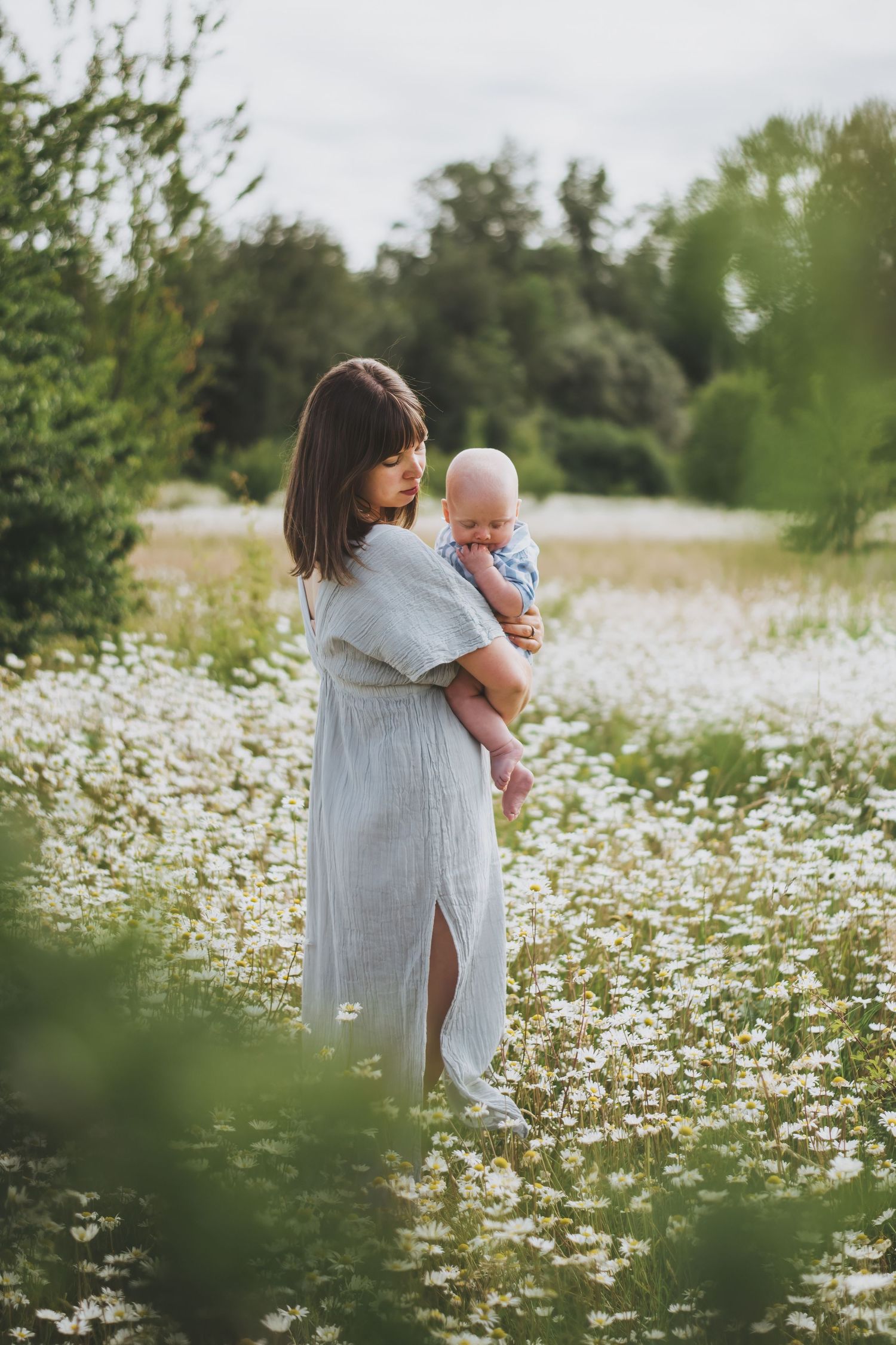 Woman in a light summer dress holds her baby while standing in a blooming field of white daisies on a bright summer day.