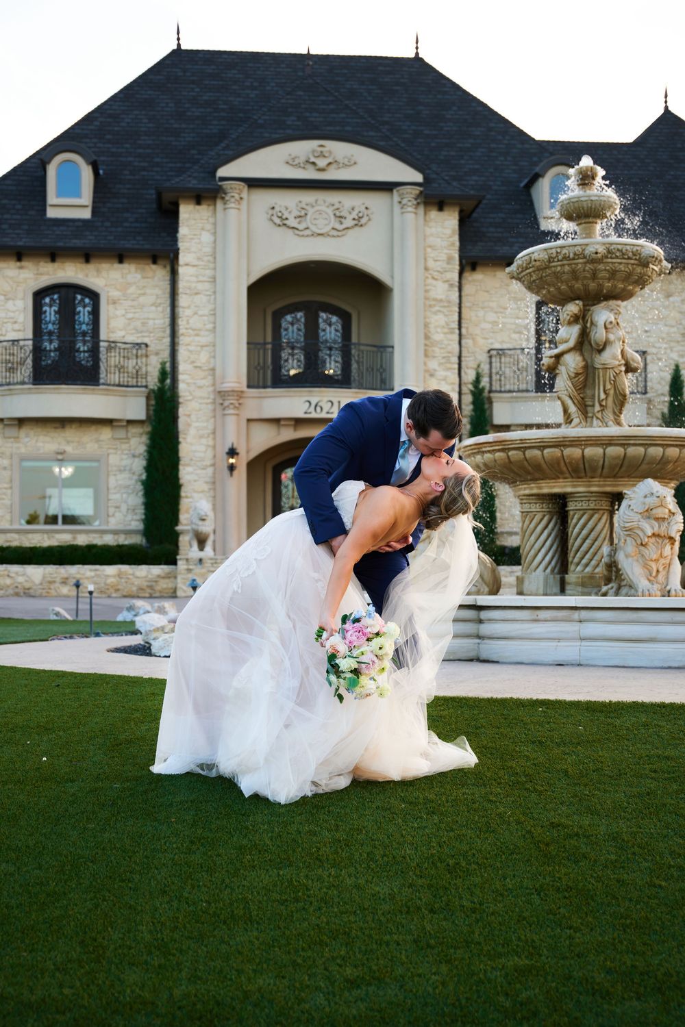 Romantic wedding couple embracing in front of a grand mansion with ornate fountain.