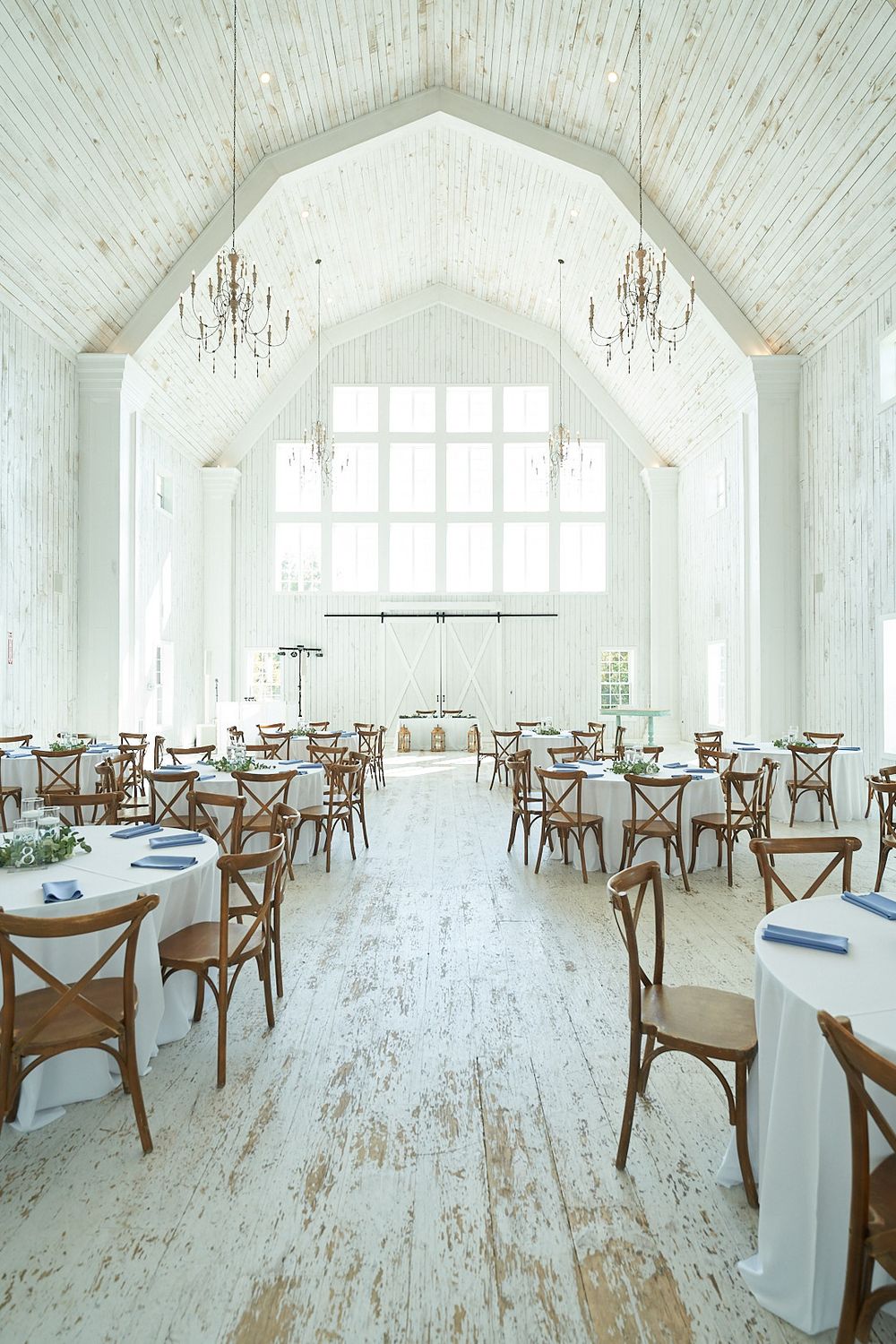 Bright white reception venue with rustic wood floors, crystal chandeliers and cross-back chairs.