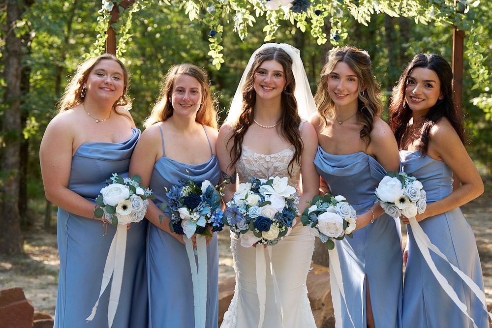 A wedding party poses with bridesmaids in light blue dresses holding white and blue floral bouquets.
