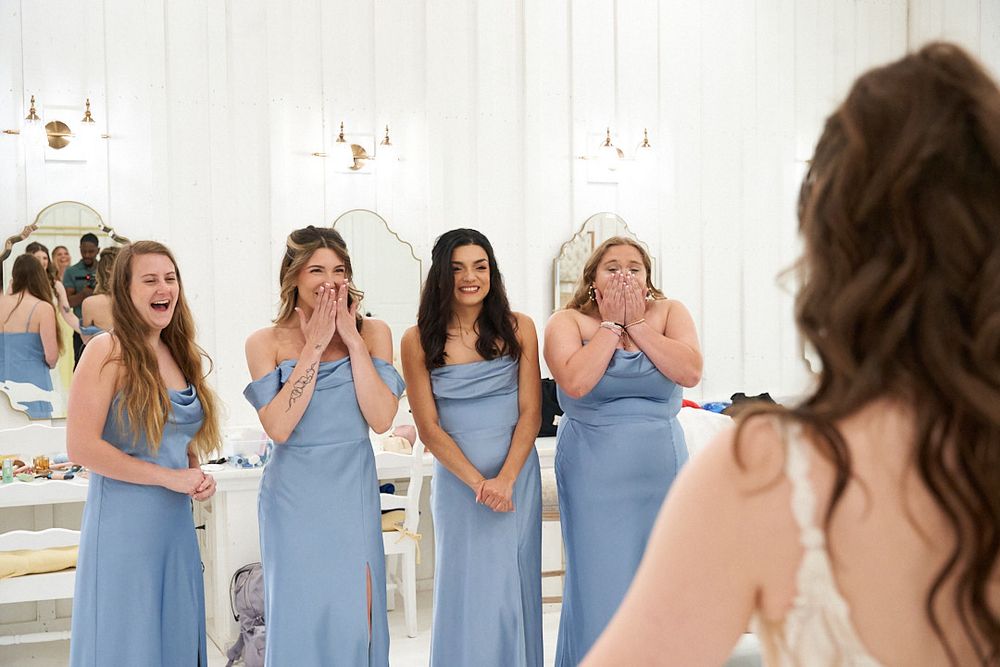 Bridesmaids in light blue dresses react with joy during wedding preparation.