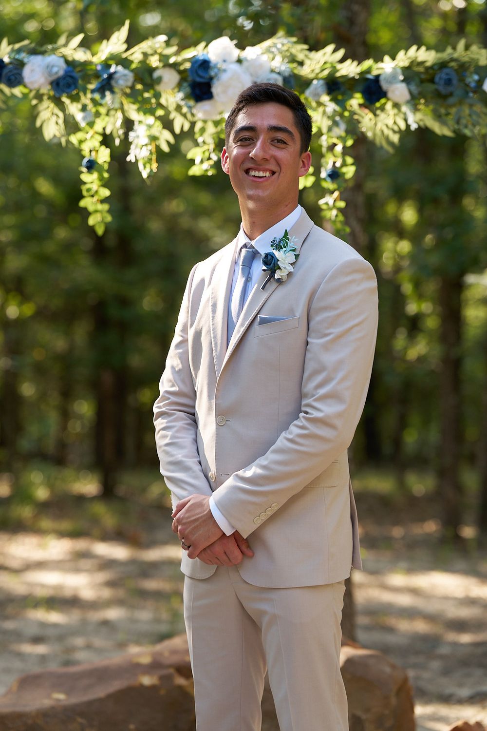 A groomsman in a light beige suit poses outdoors under green foliage at a wedding.