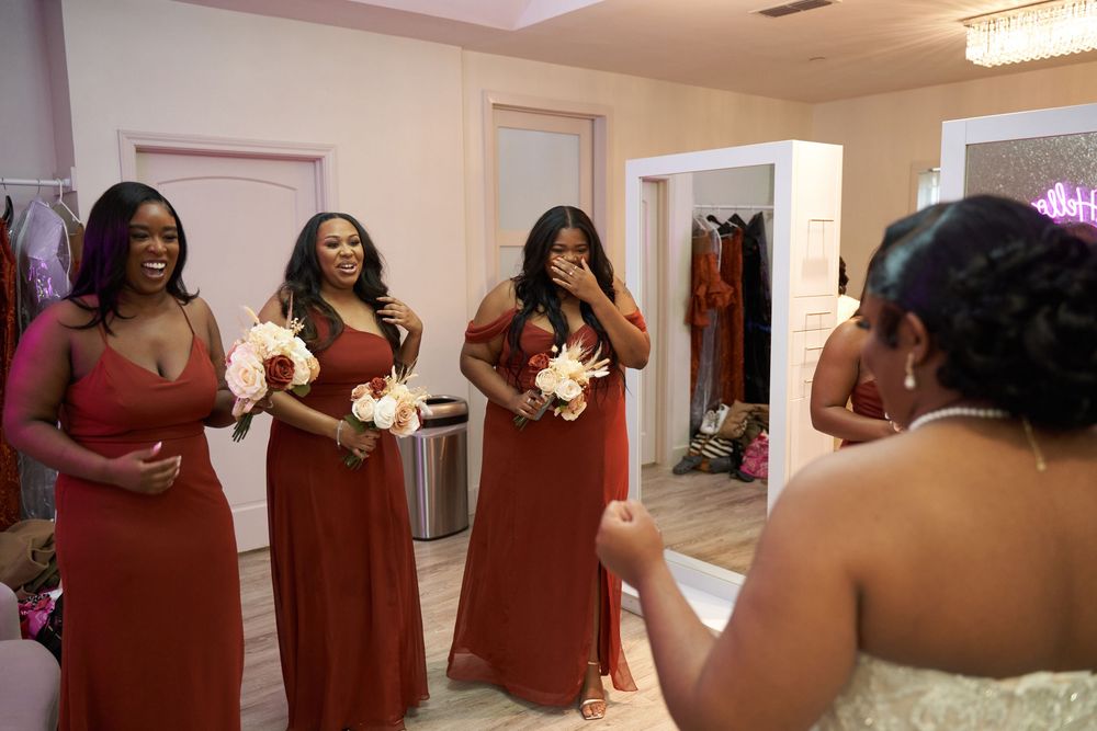 Bridesmaids in burgundy dresses share excited reactions in dressing room.