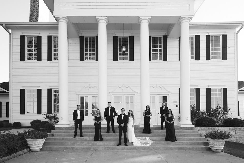 A formal wedding party poses in front of a white columned mansion with classic architecture.