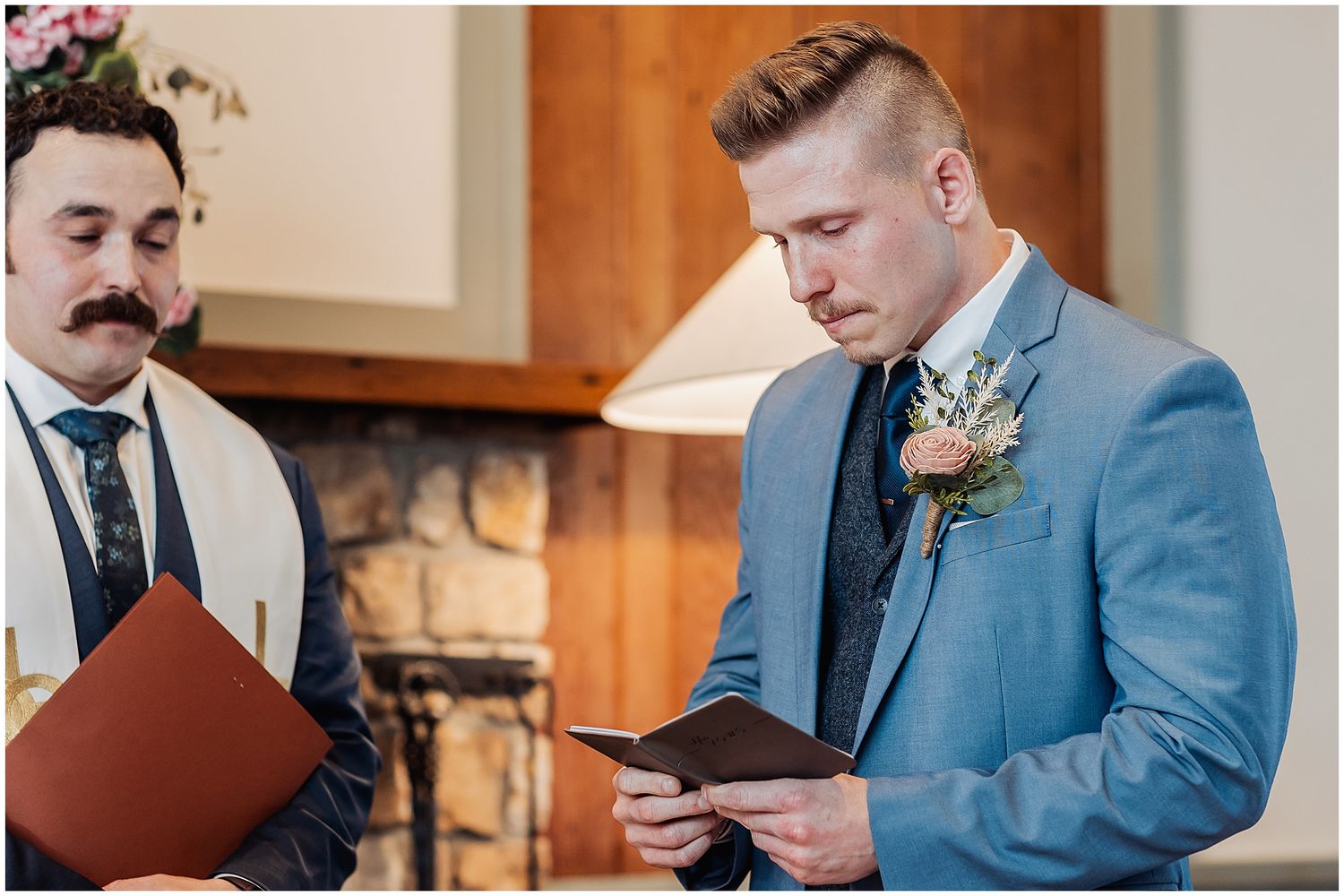 A wedding ceremony moment as a groom in a blue suit reads vows from a tablet while an officiant looks on.