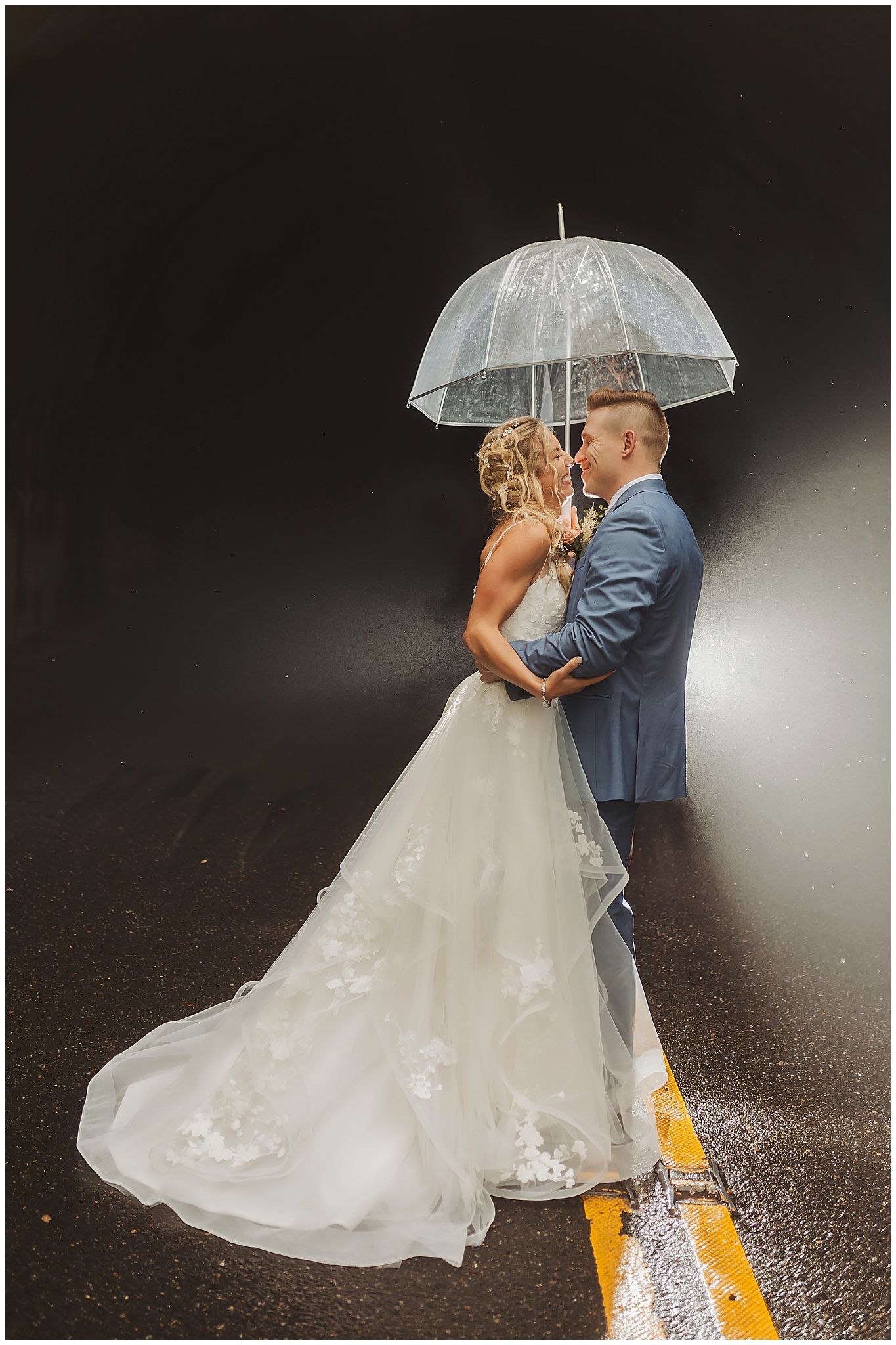 Romantic rainy wedding photo with couple under clear umbrella on wet road with yellow line.