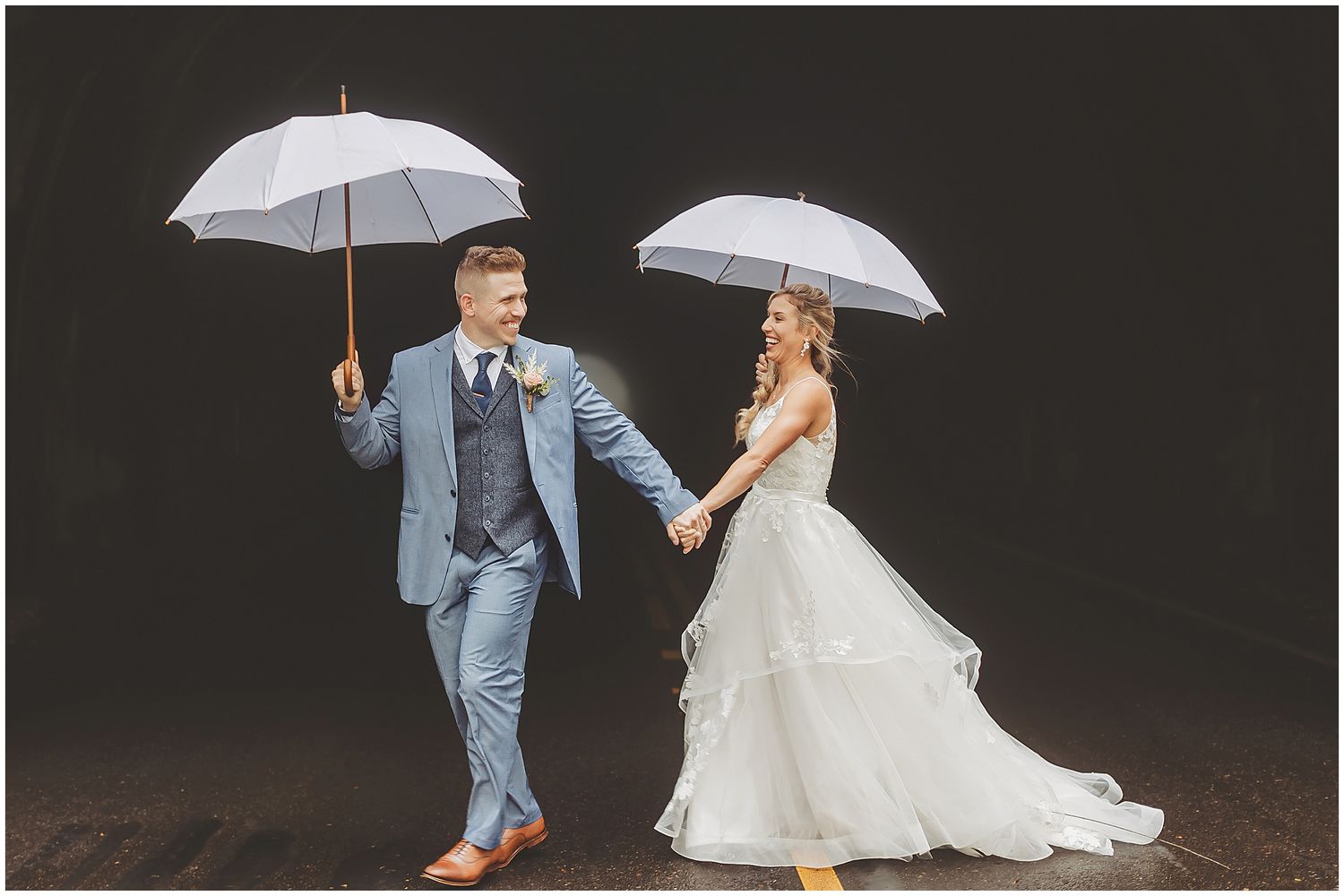 Newlyweds with white umbrellas walk together against dark background in stylish wedding attire.