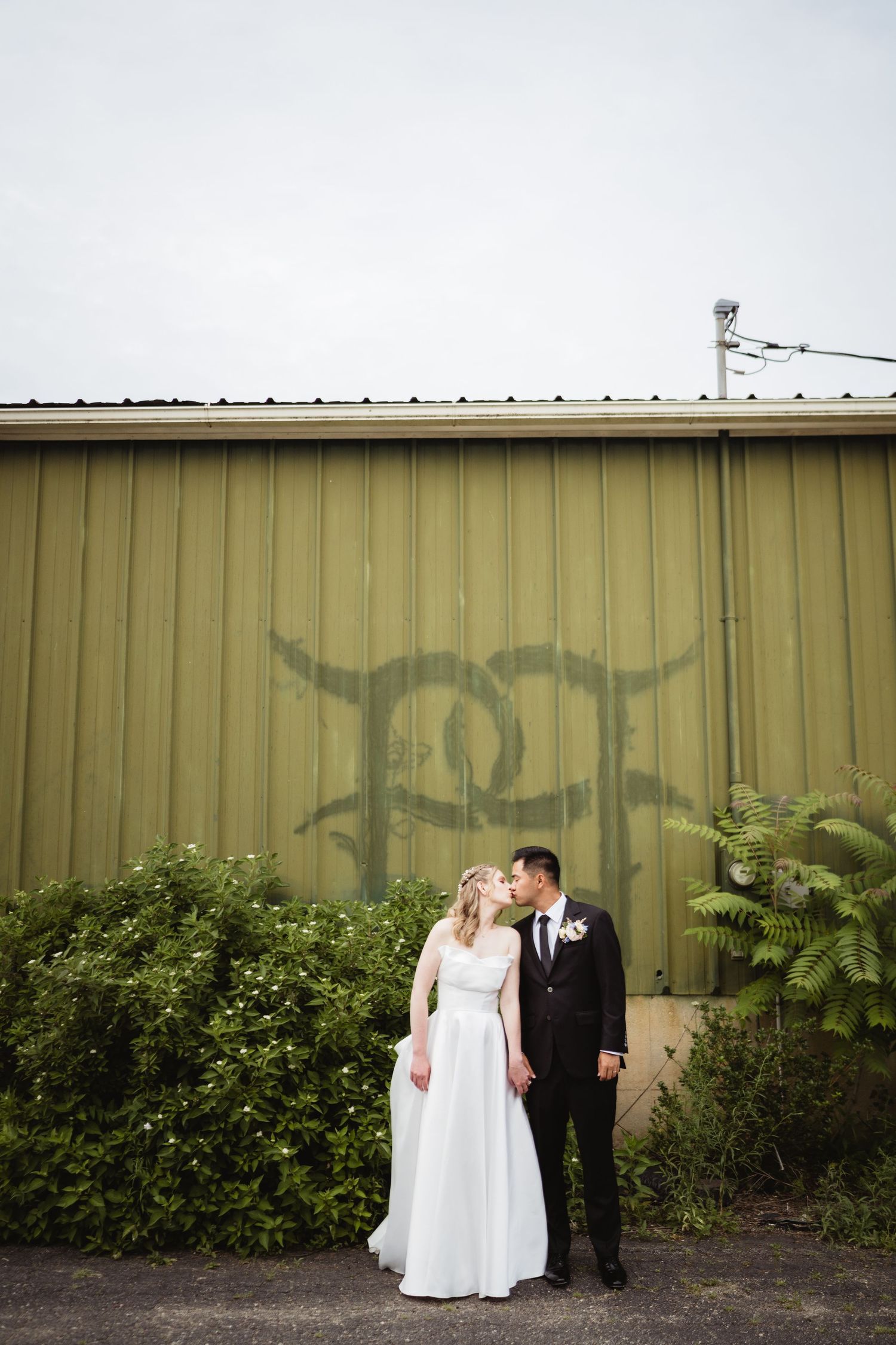 A couple in wedding attire poses in front of an olive green metal wall with greenery and a painted design.
